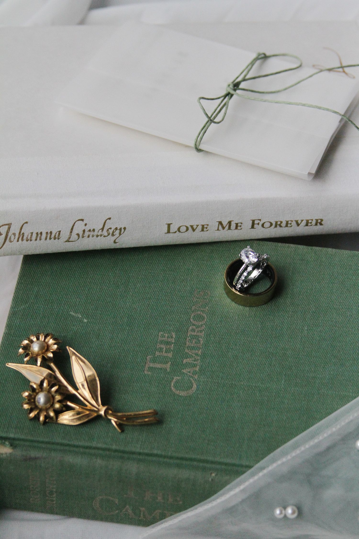Vintage wedding flat lay with diamond rings, gold brooch, and Johanna Lindsey romance novel on green book.