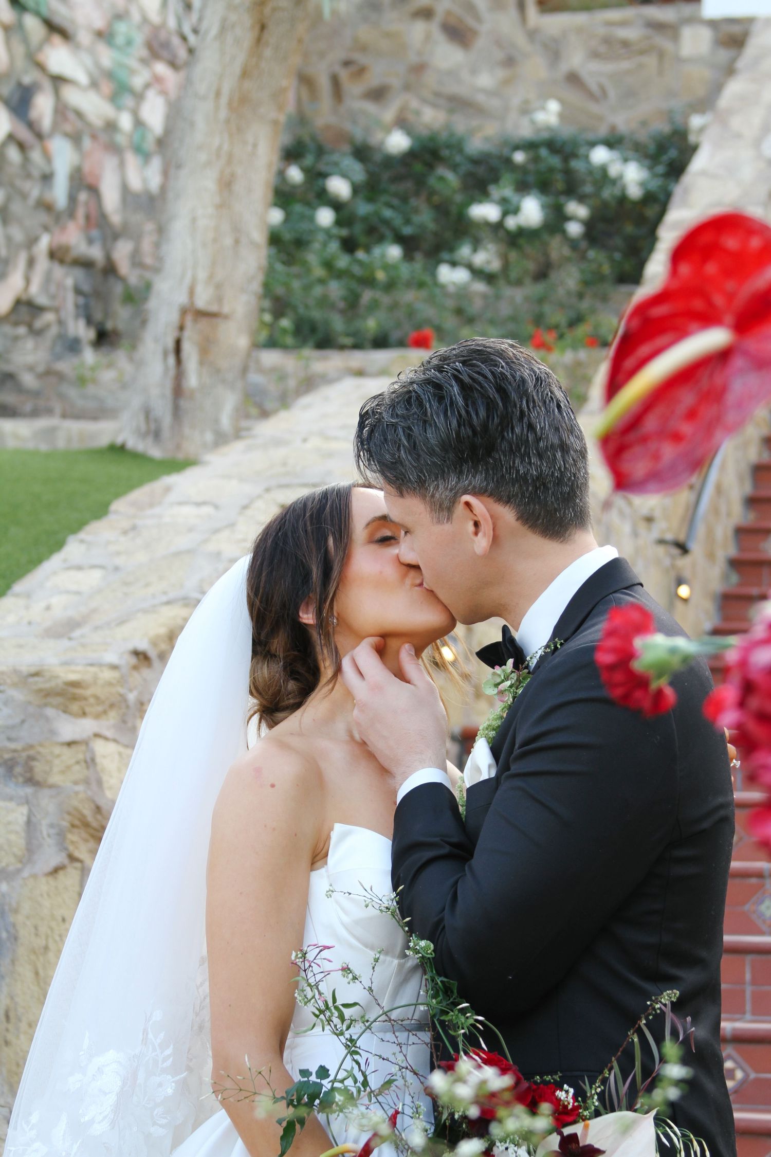 Bride and groom share a romantic first kiss on stone steps surrounded by red flowers at their outdoor wedding.