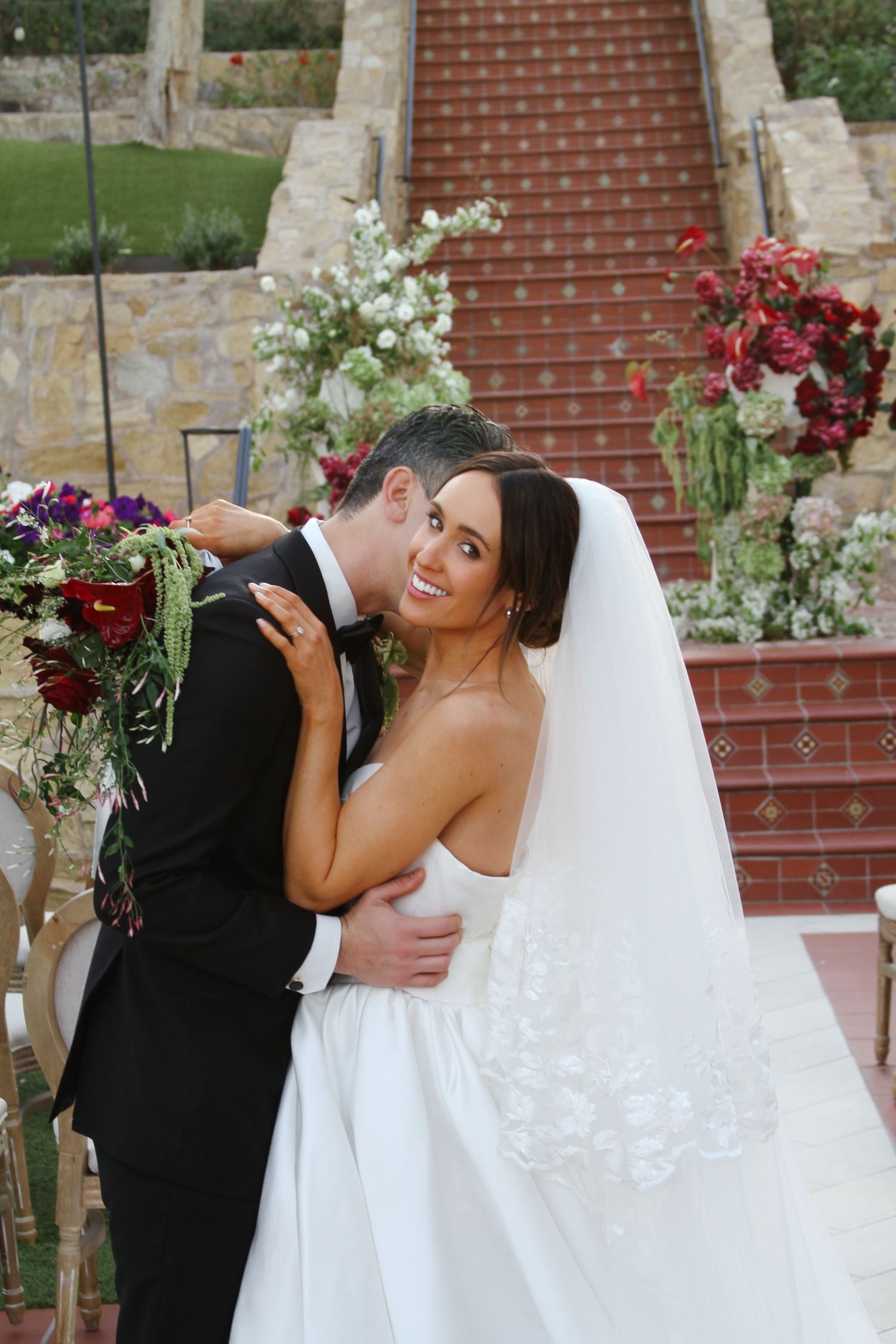 Bride in white strapless gown and veil holds bouquet while groom in black tuxedo embraces her on elegant outdoor wedding venue steps.