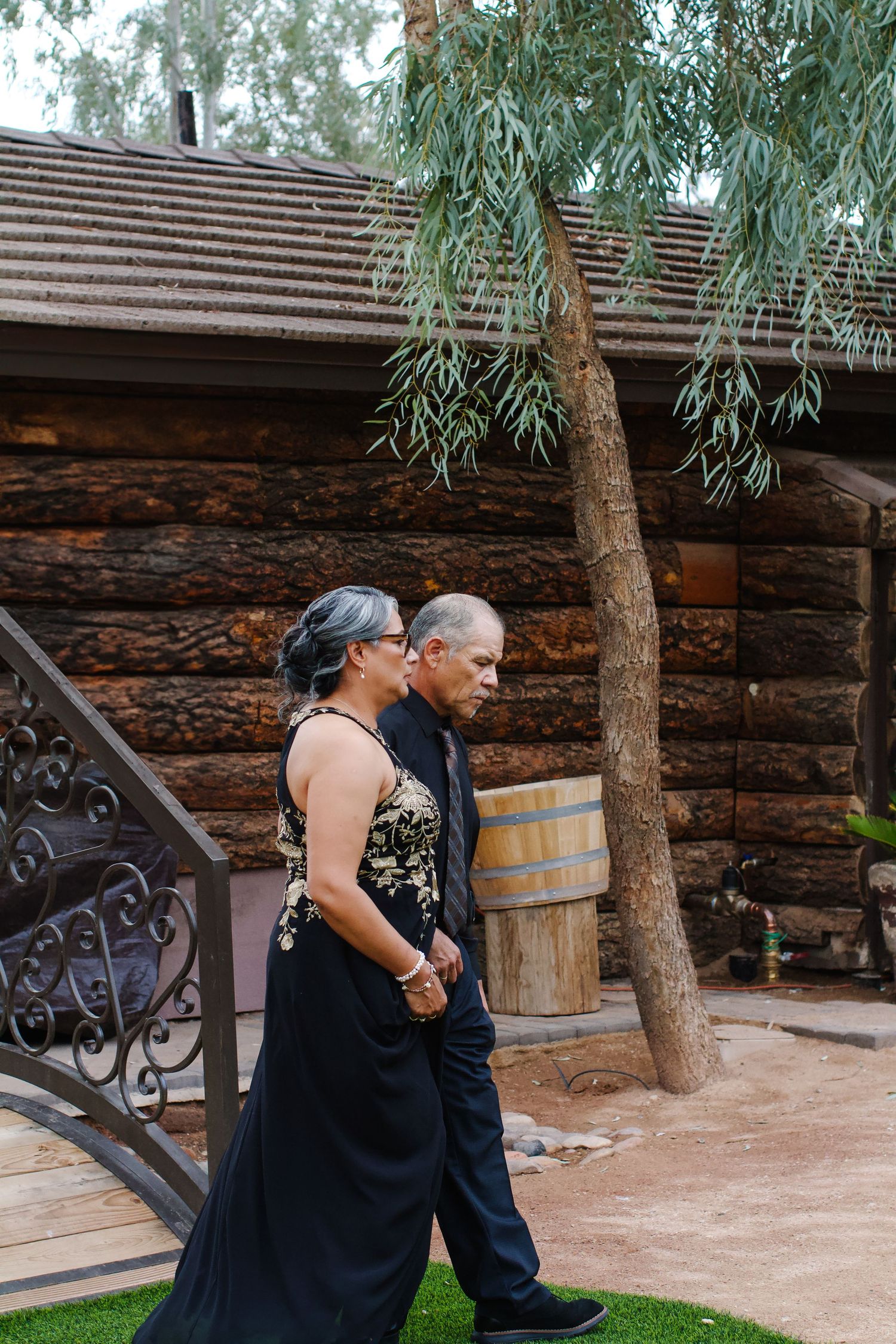 Two people walk together near a rustic log cabin with a willow tree in the foreground.