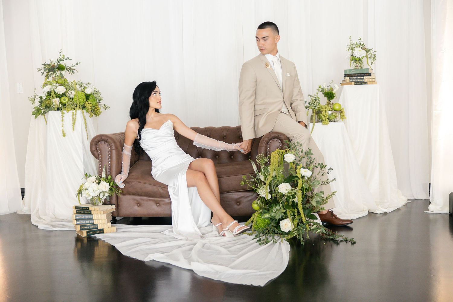 Bride in white gown sits on brown leather sofa beside groom in tan suit, surrounded by white and green floral arrangements.