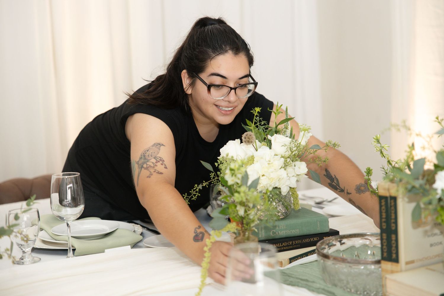 Woman with tattoos arranging white floral centerpiece on elegant table setting with glassware and green books.