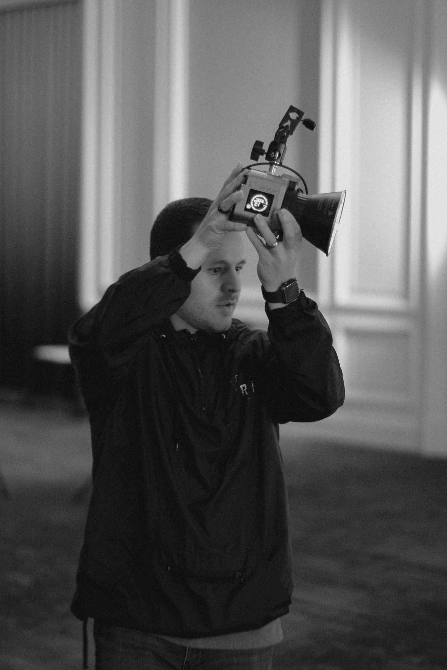 Black and white photo of a man holding a professional video camera above his head in an indoor setting.