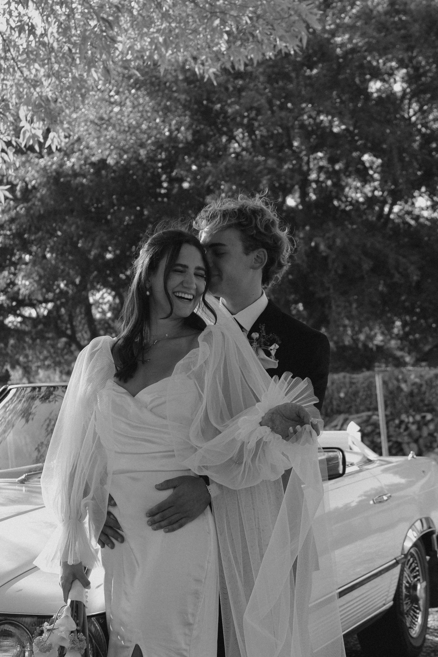 A romantic black and white wedding photo of a couple sharing a joyful moment in front of a vintage car.