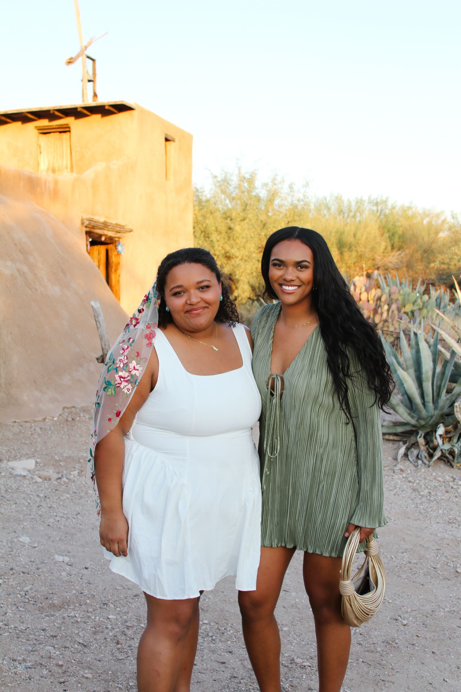 Two friends stand together in desert setting with white and green dresses against adobe wall and agave plants.