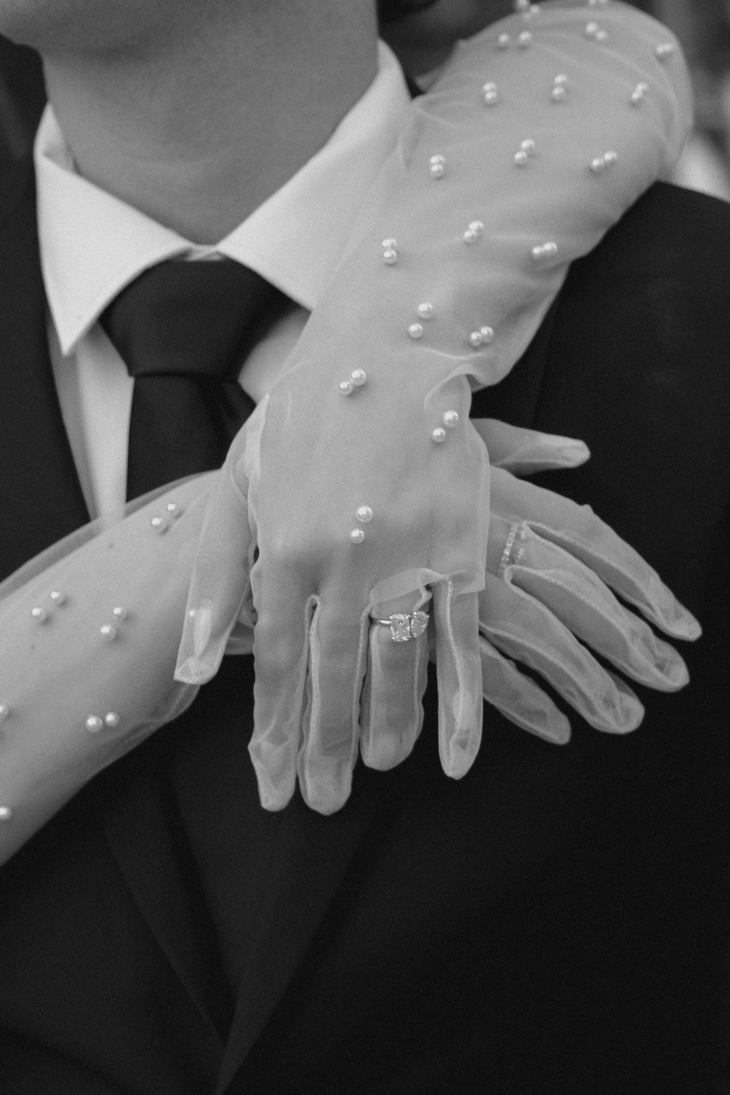 Black and white photo of bride wearing pearl-embellished gloves and engagement ring, hands resting on groom's suit.