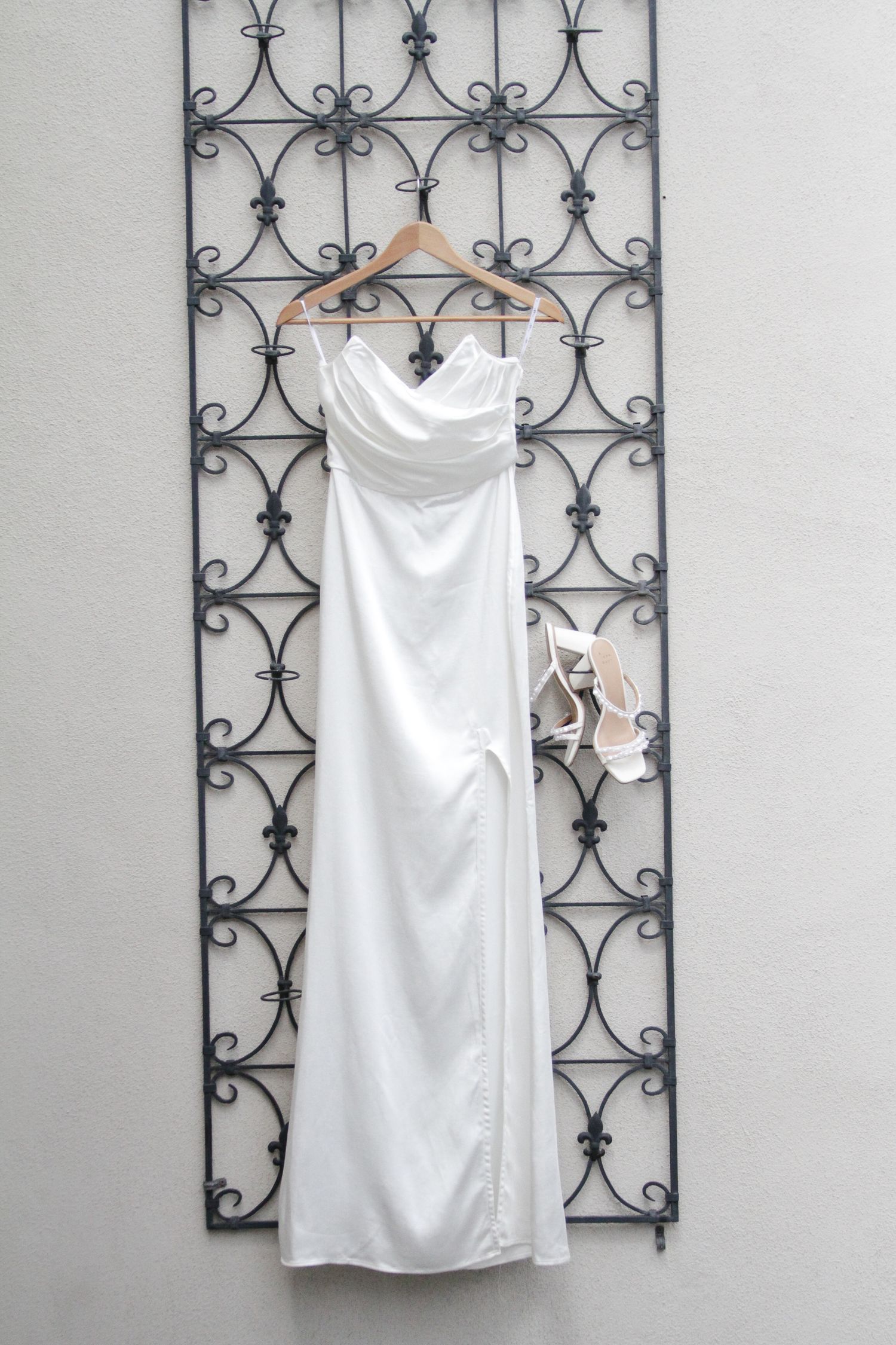 Minimalist white satin wedding gown with cowl neckline hanging on wooden hanger with strappy heels.