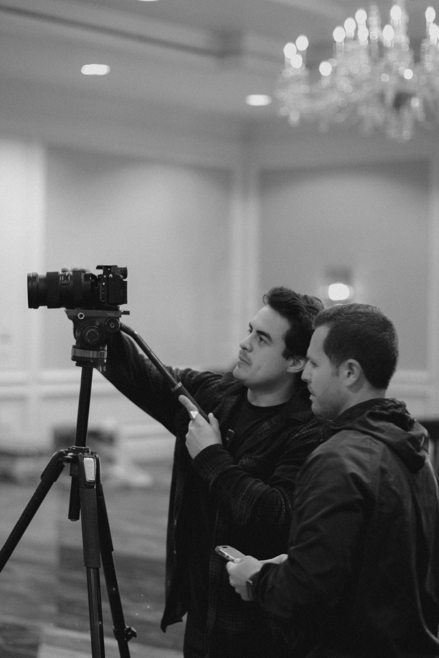 Two men in black jackets adjust a camera on a tripod in a ballroom, captured in black and white photography.