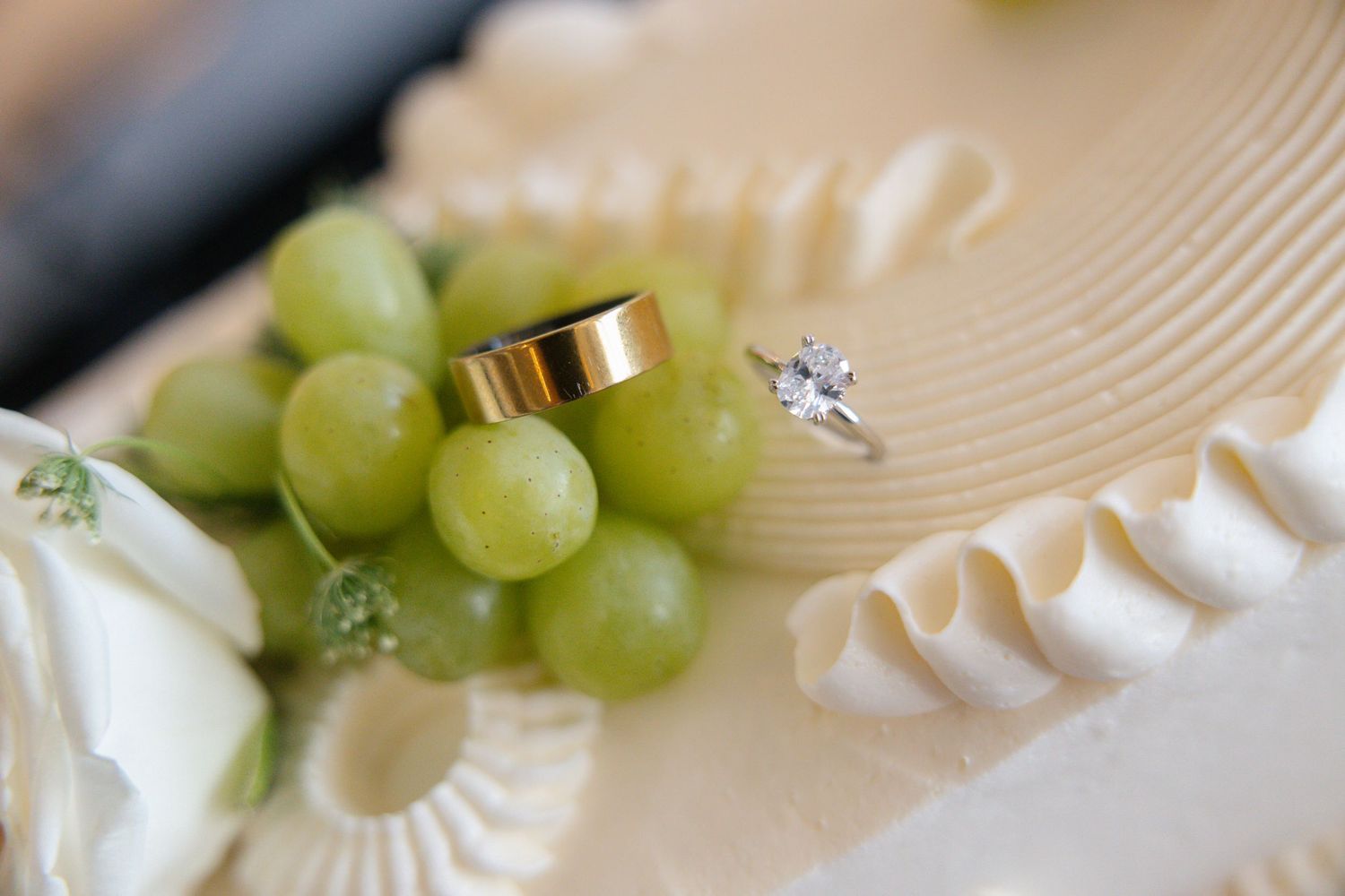 Gold wedding band and diamond engagement ring resting on green grapes atop a white wedding cake.