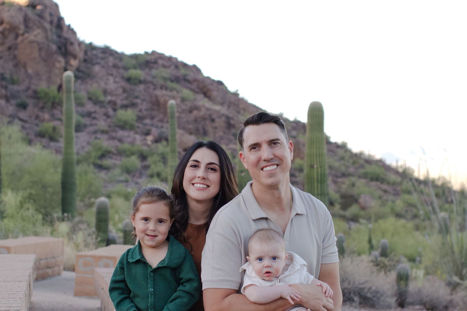 Family poses together on desert trail with saguaro cactus and rocky mountains in background during sunset.