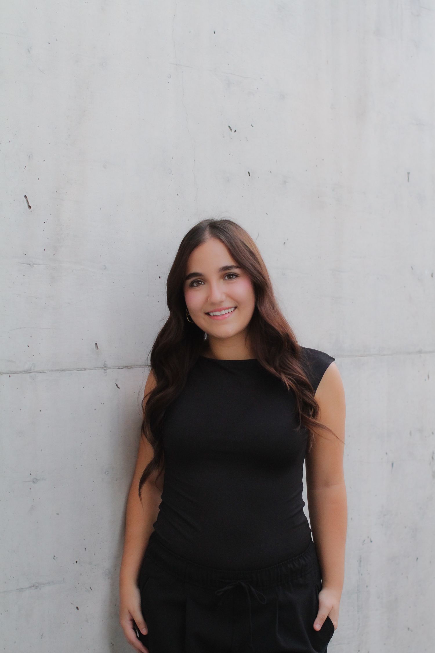 Woman in a black sleeveless top smiles while posing against a light colored wall.