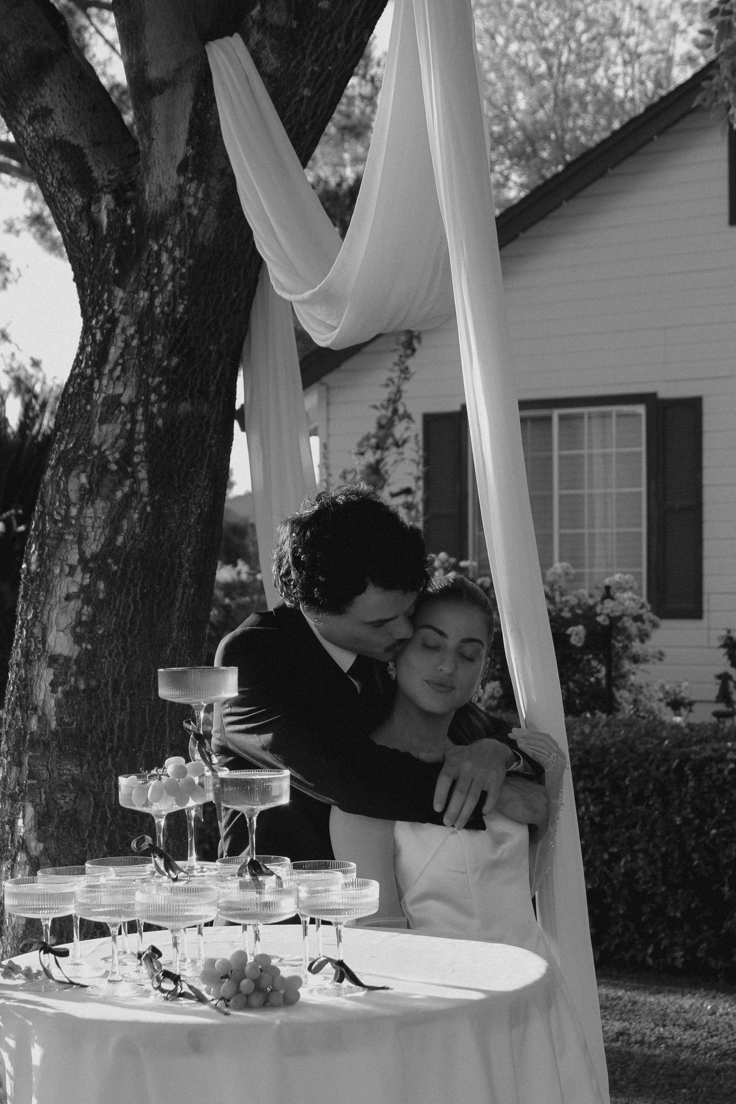 Black and white photo of a bride and groom embracing near a champagne tower at an outdoor wedding reception.