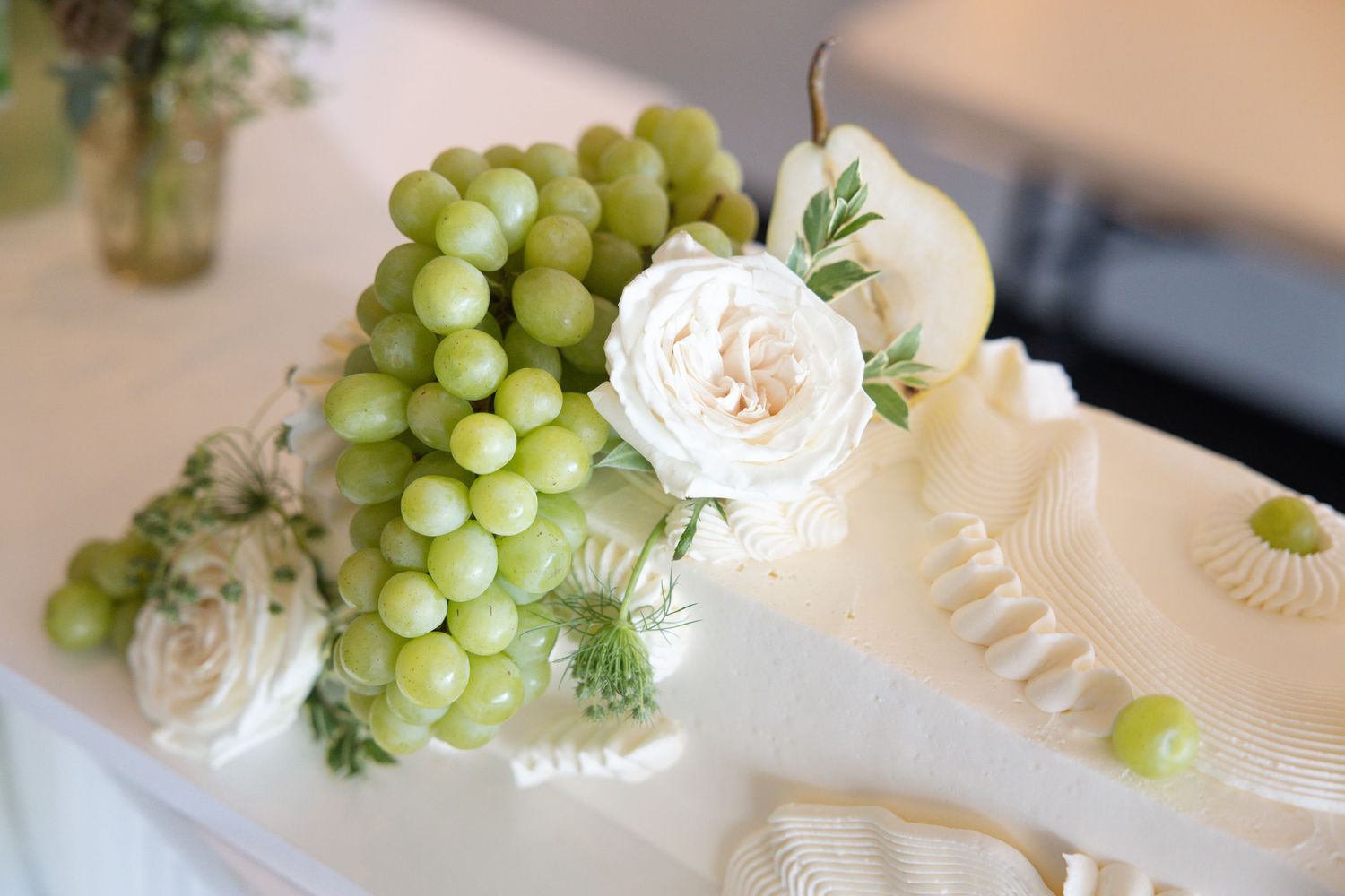 White wedding cake adorned with green grapes, a pear, and white roses on a elegant table setting.