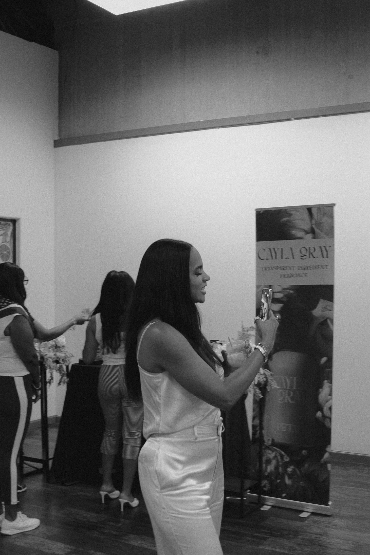 Woman in white outfit photographing a Cayla Gray art exhibition banner in a gallery space.
