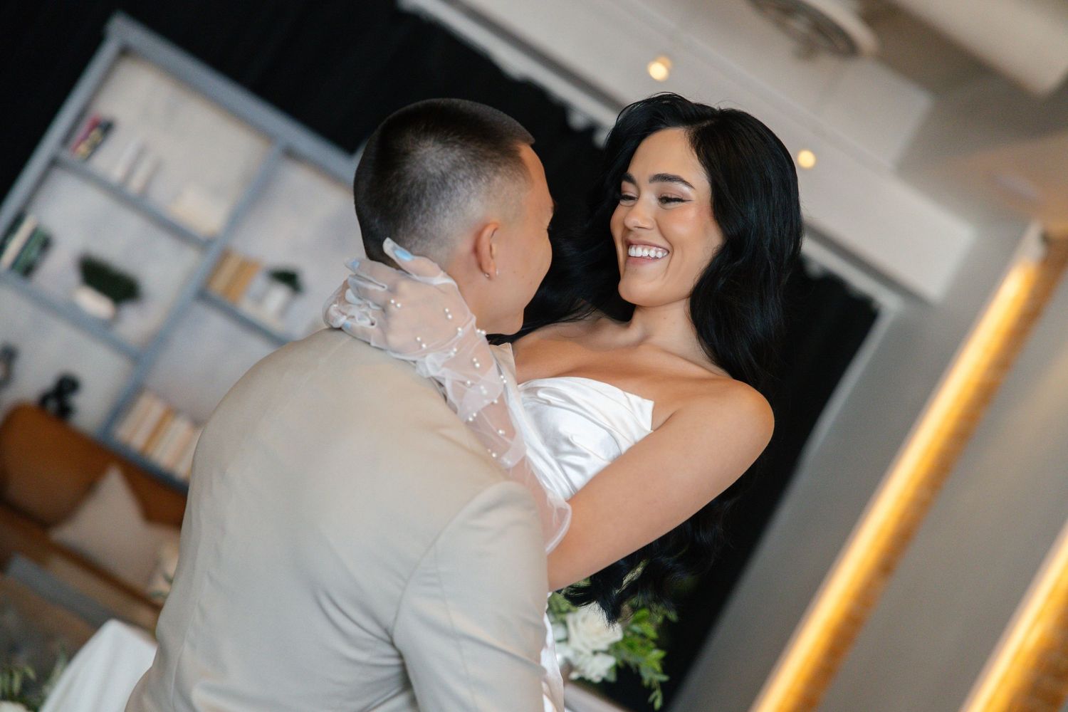 Joyful bride in strapless white gown shares first dance with groom in beige suit.