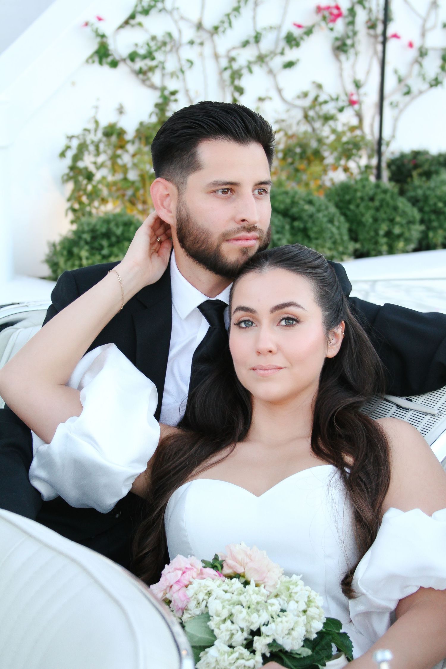 A couple in wedding attire poses on a white couch with pink and white flowers in the background.