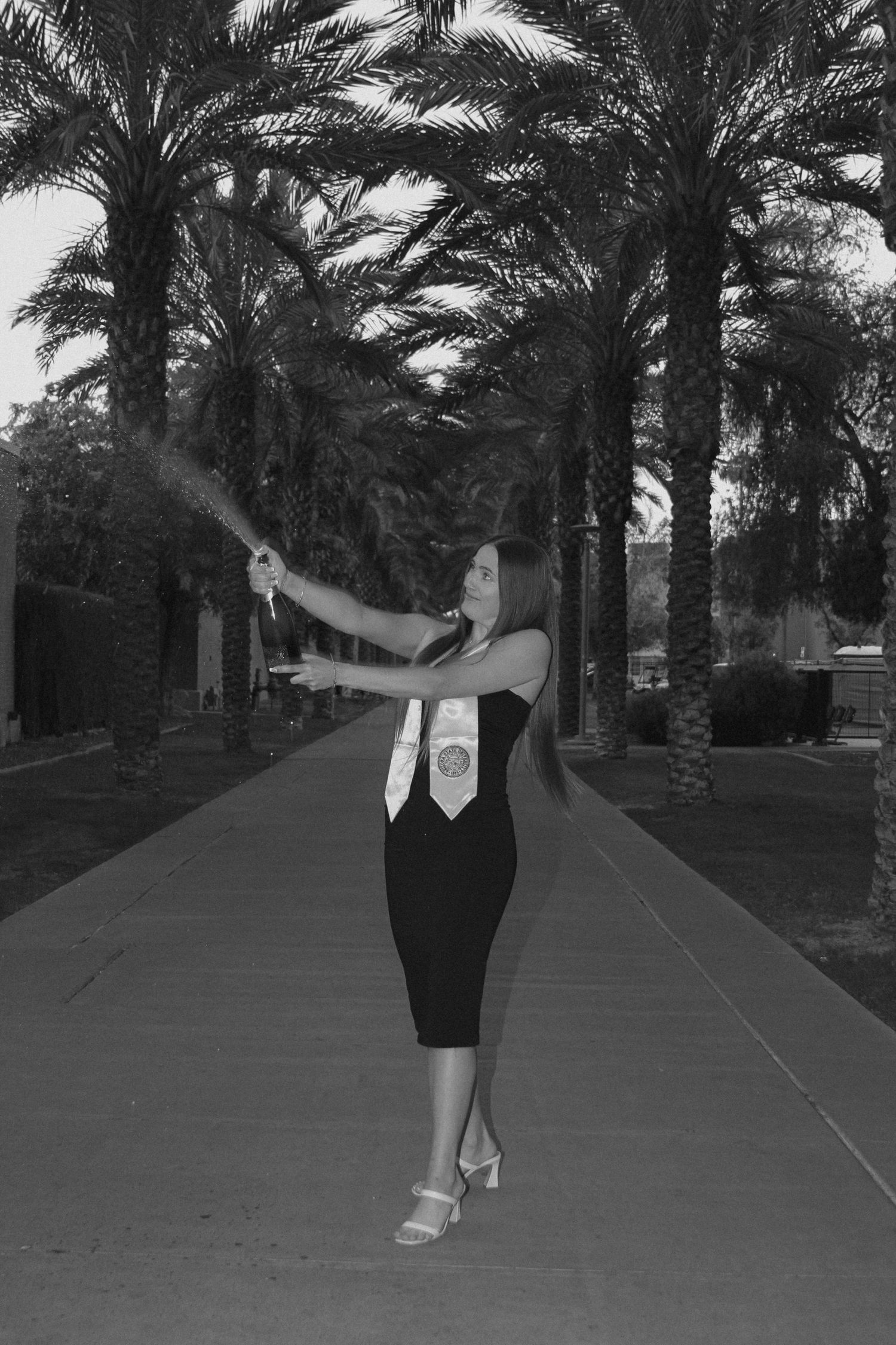 A graduate in a black dress celebrates with sparklers along a palm tree-lined path at night.