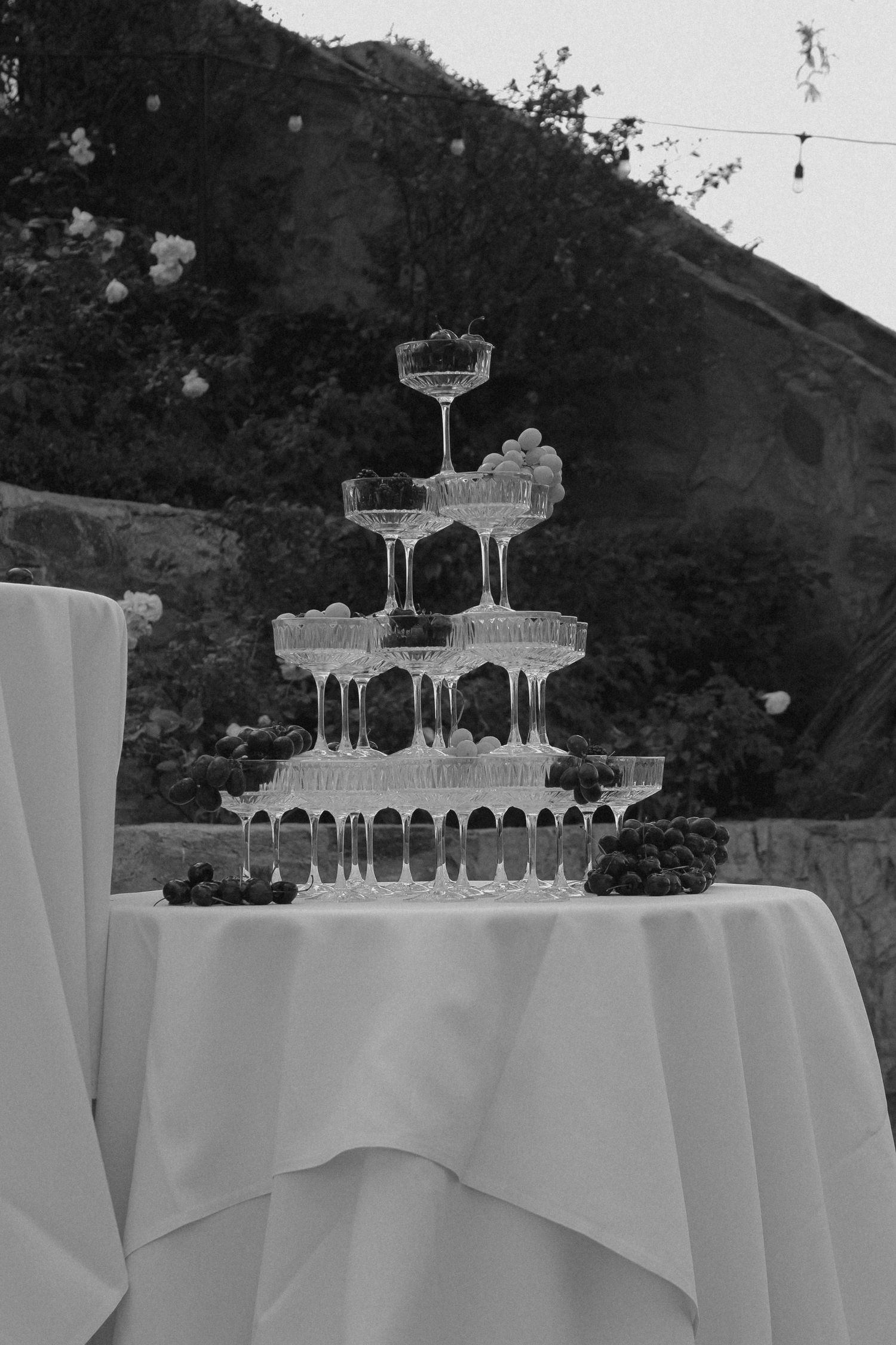 Black and white photo of a champagne tower with grapes on a white draped table outdoors.