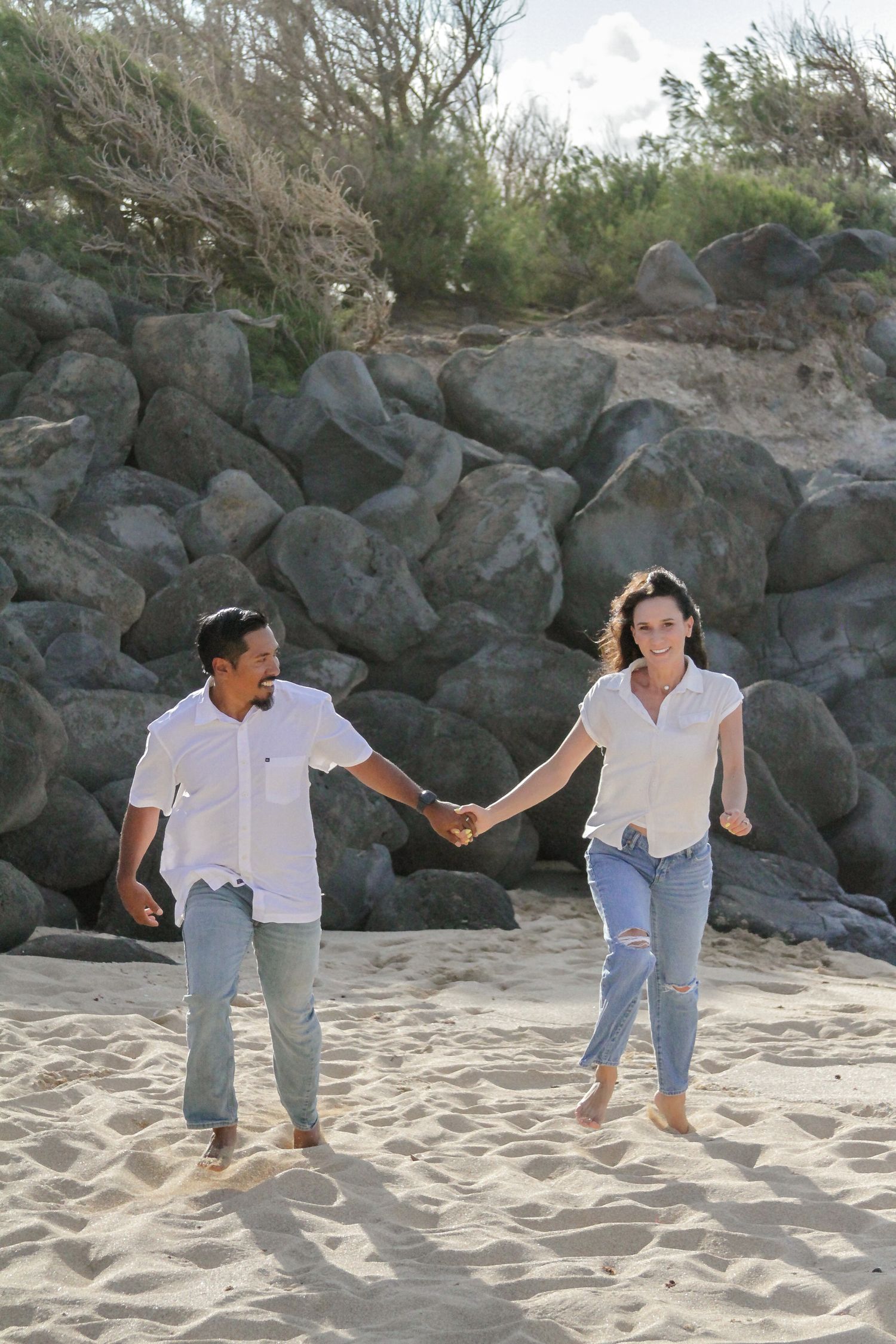 A couple holds hands while walking barefoot on a sandy beach with large rocks and greenery in the background.
