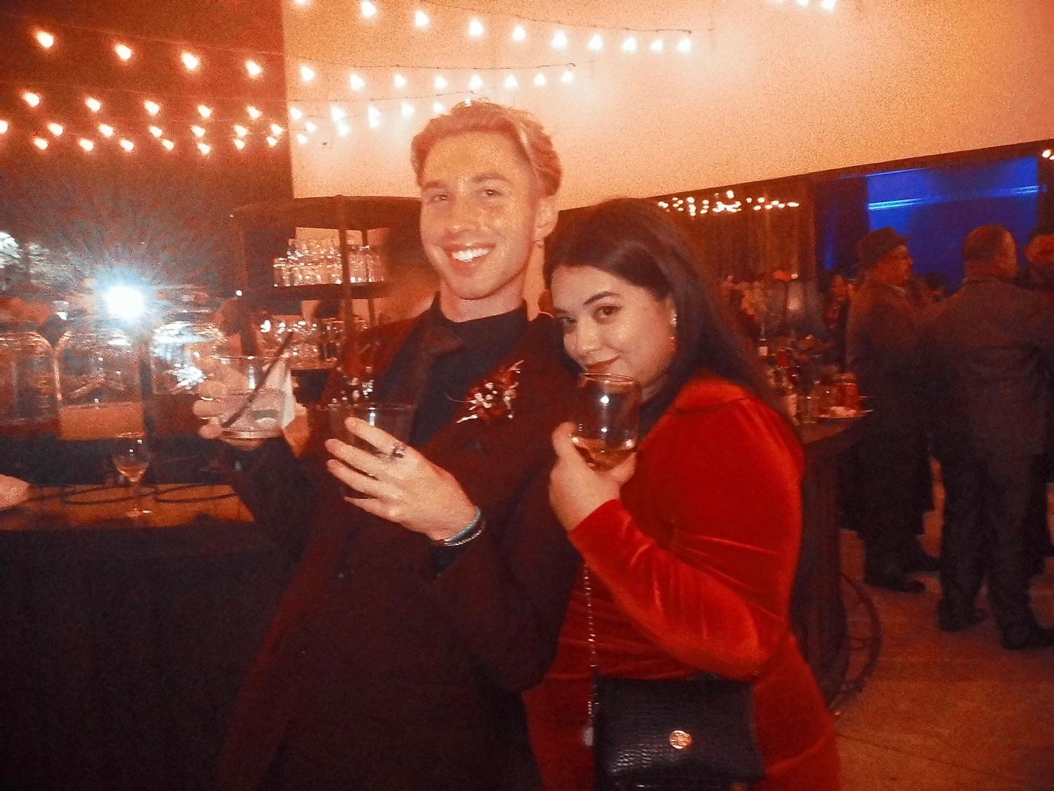 Smiling couple in formal attire holding cocktails at a warmly lit indoor wedding reception venue.