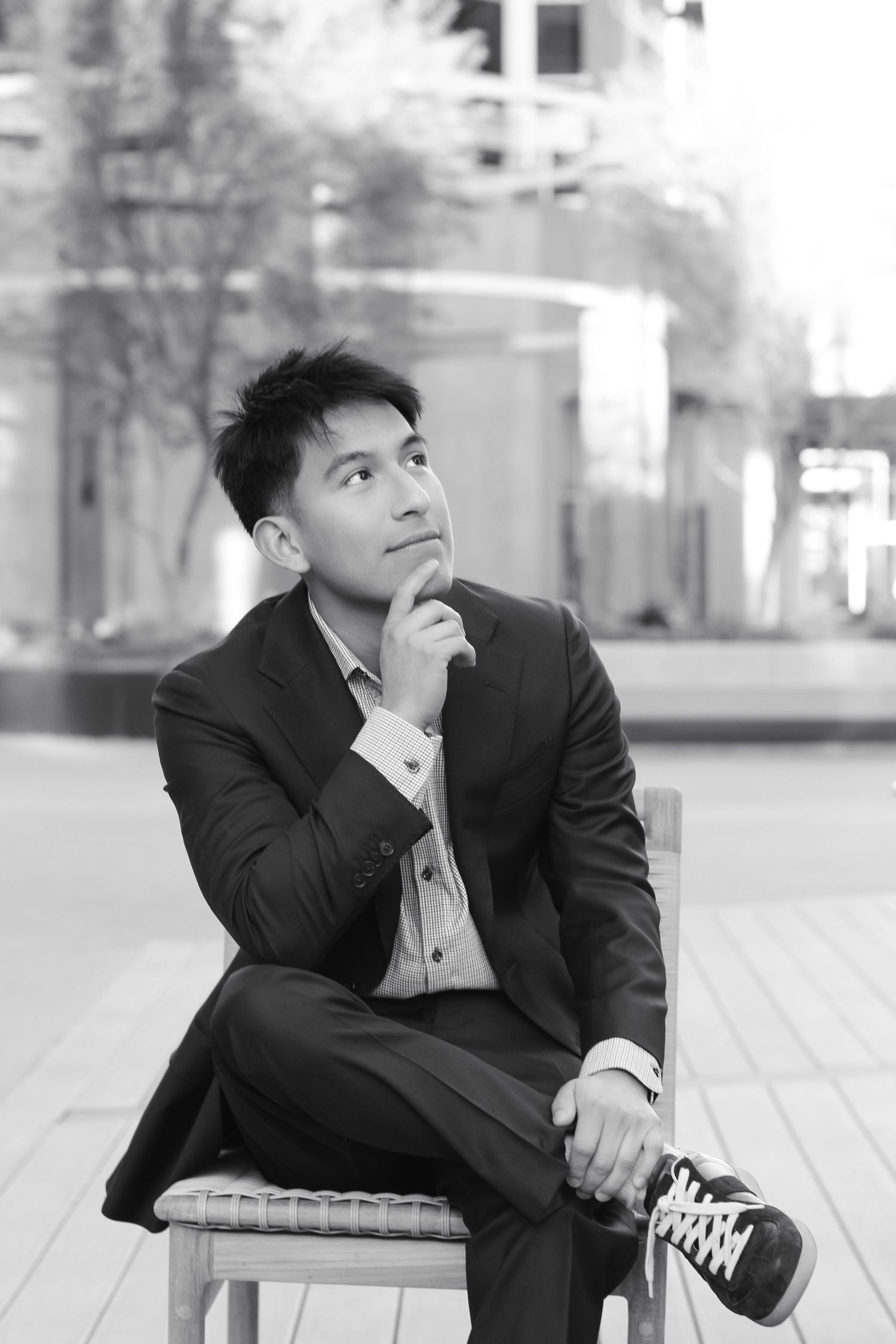 Black and white portrait of person in business attire sitting thoughtfully on chair outdoors.