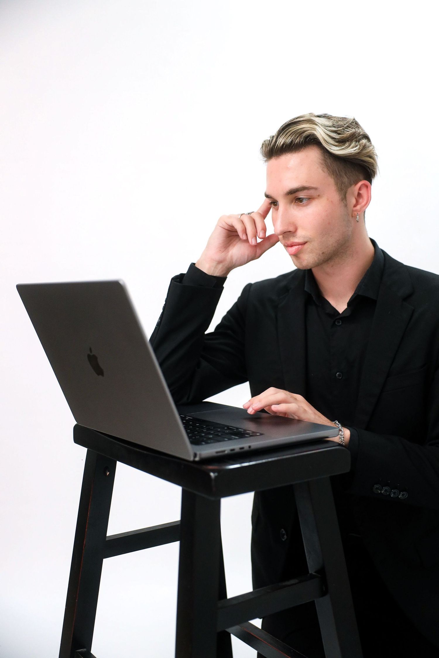 Young man in black suit working on Apple MacBook laptop, resting chin on hand in thoughtful pose against white background.