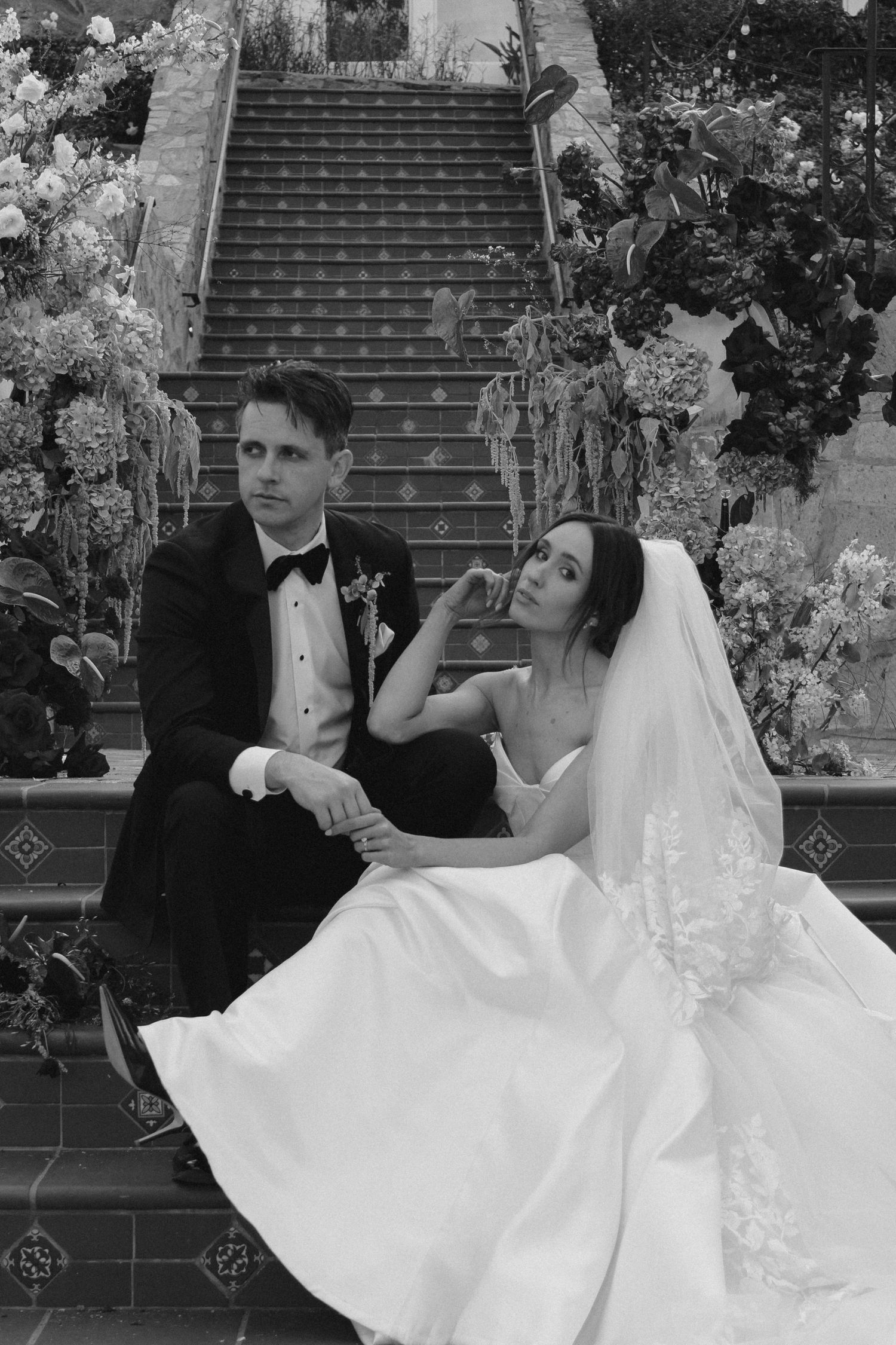 Black and white photo of bride and groom sitting on tiled steps surrounded by floral arrangements.