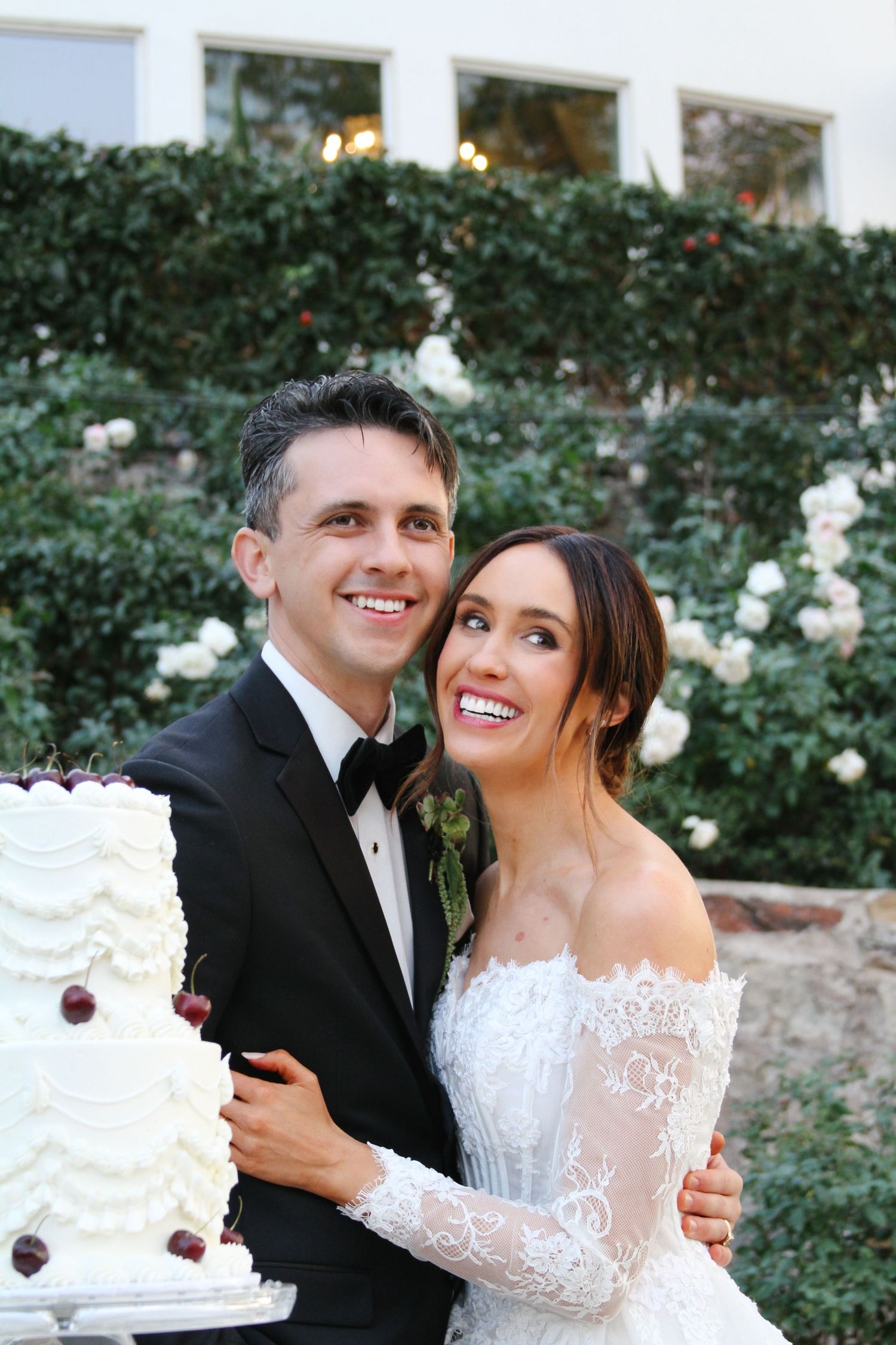 Smiling bride and groom pose together with elegant white tiered wedding cake decorated with dark cherries outdoors.