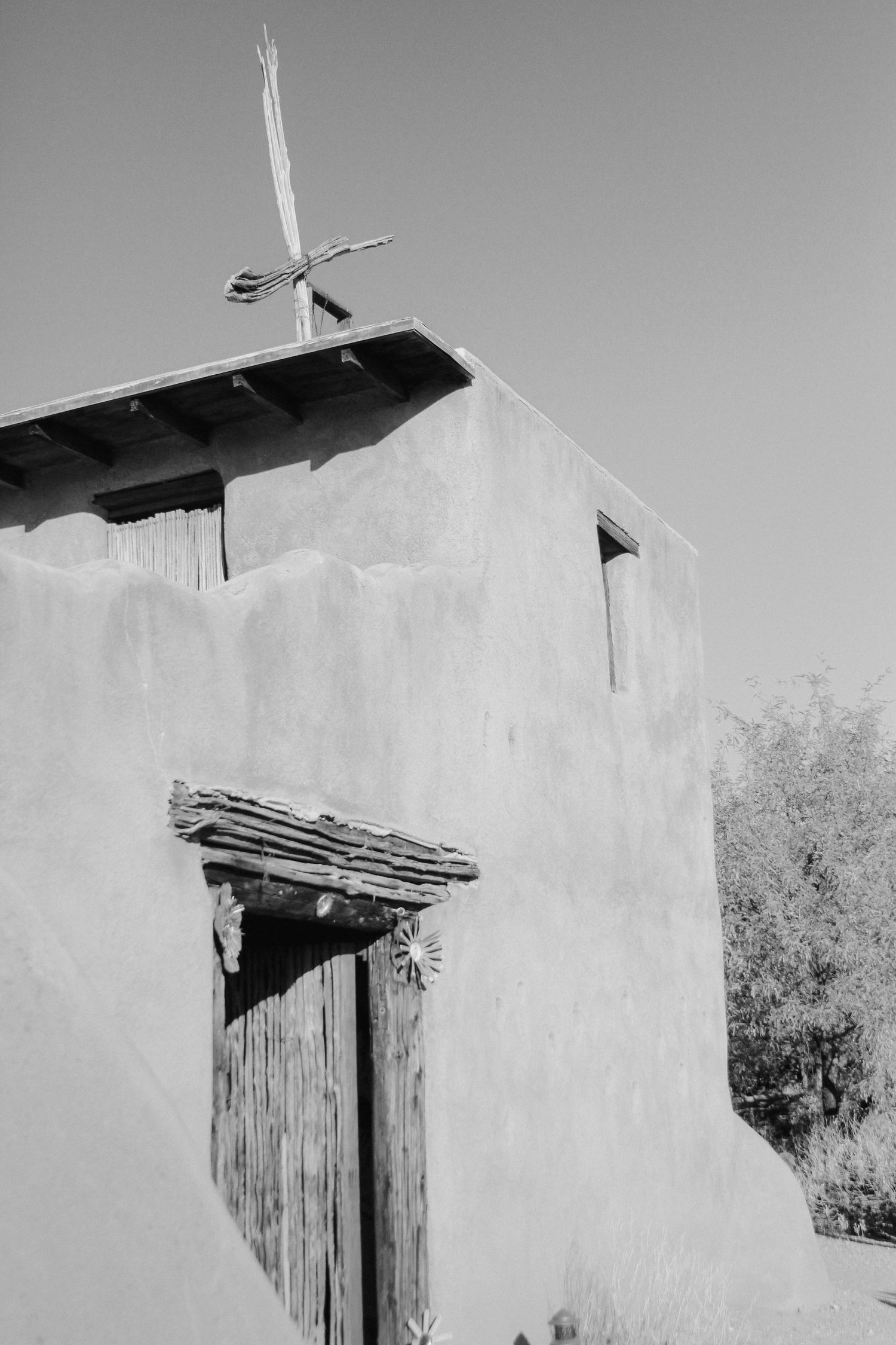 Black and white photo of rustic adobe chapel with wooden door and cross on roof.