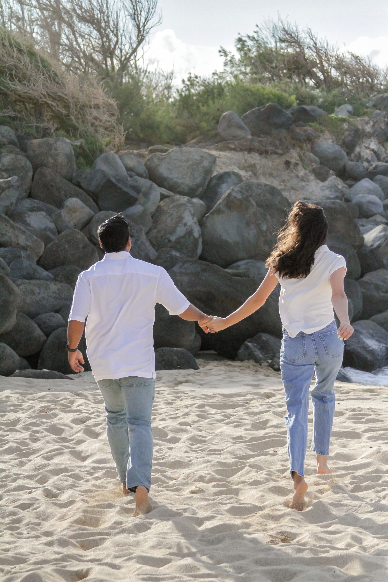 Two people walking hand in hand along a sandy beach with rocky formations in the background.
