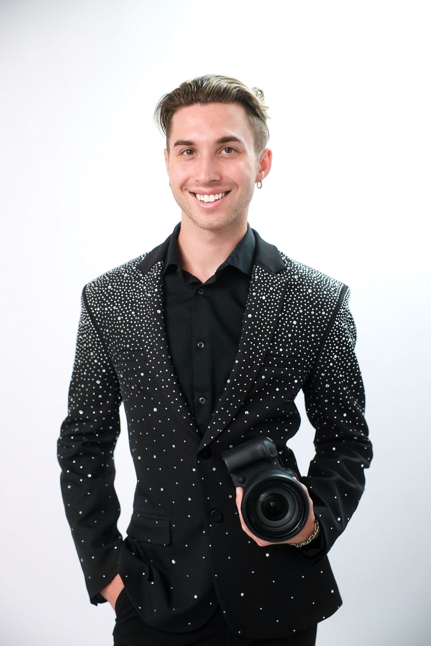 Smiling young man in a black studded blazer holding a professional camera against a white background.
