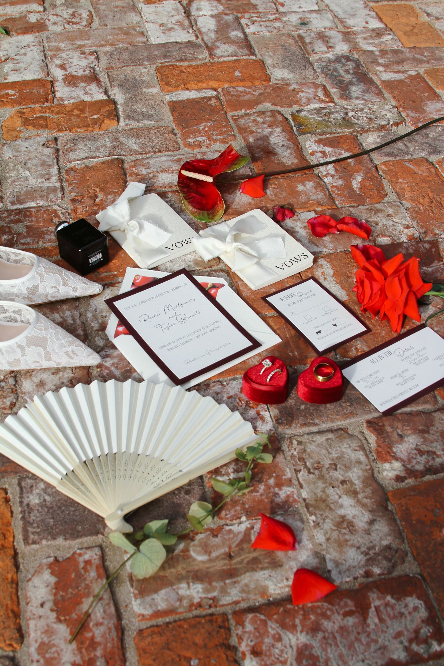 Wedding stationery, red rose petals, a hand fan, and ring boxes arranged on rustic brick pavement.