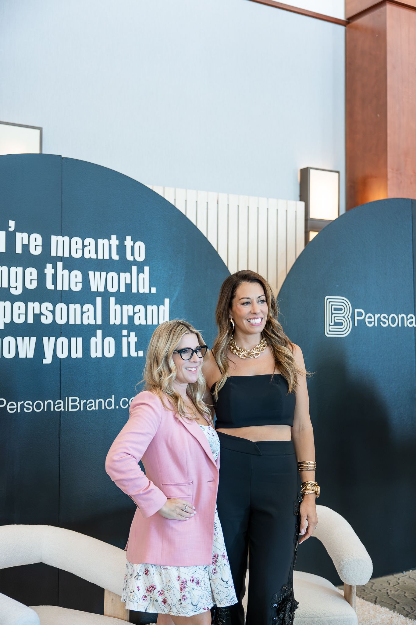 Two women smiling in front of a PersonalBrand.com motivational sign at a professional branding event.