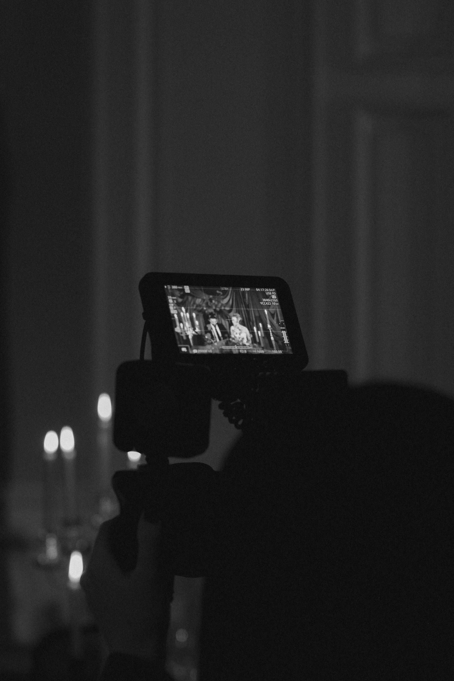 A silhouetted figure holds up a camera or device displaying video content in a dark environment with bokeh lights.