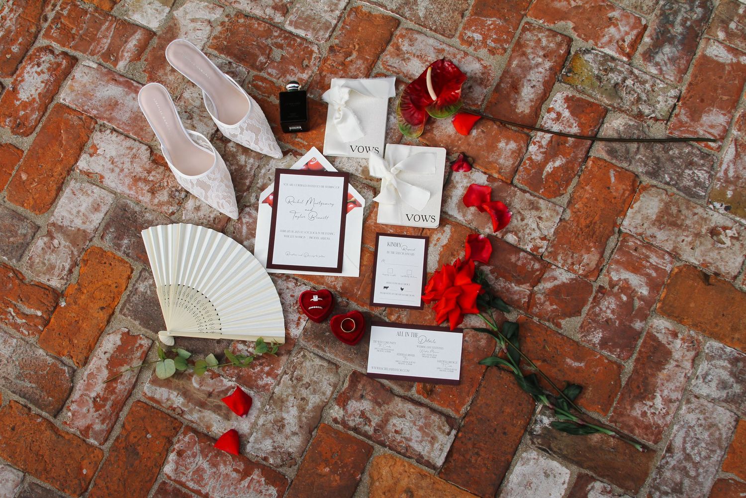 Wedding stationery flatlay with red roses and fan on rustic brick background.