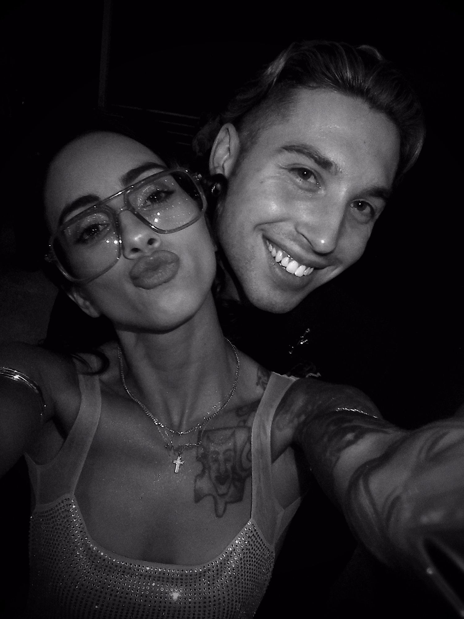 Black and white selfie of a smiling couple, woman wearing glasses and a sparkly top, man with tattoos.