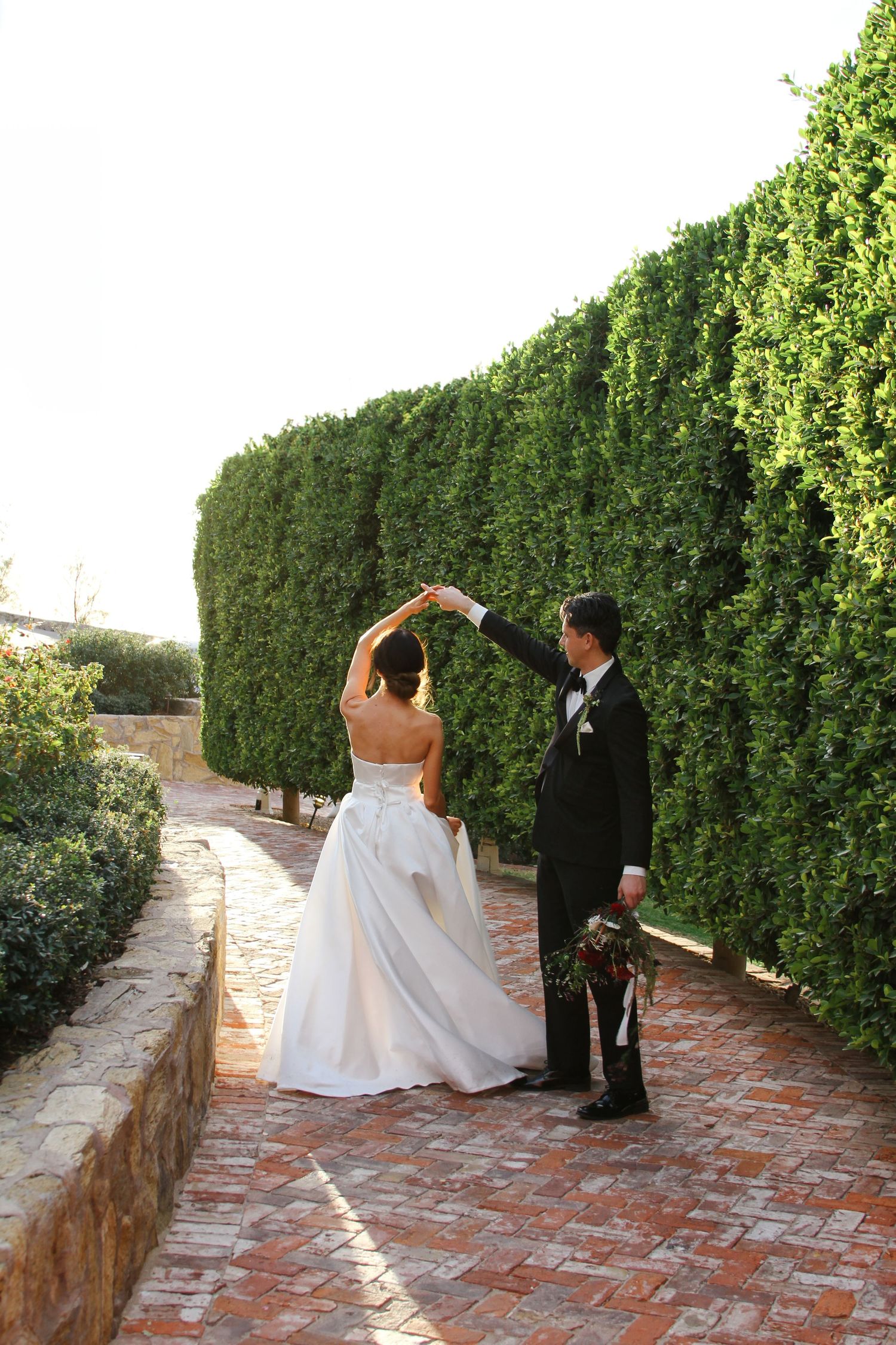 Bride and groom dancing on brick path surrounded by tall green hedges during outdoor wedding celebration.