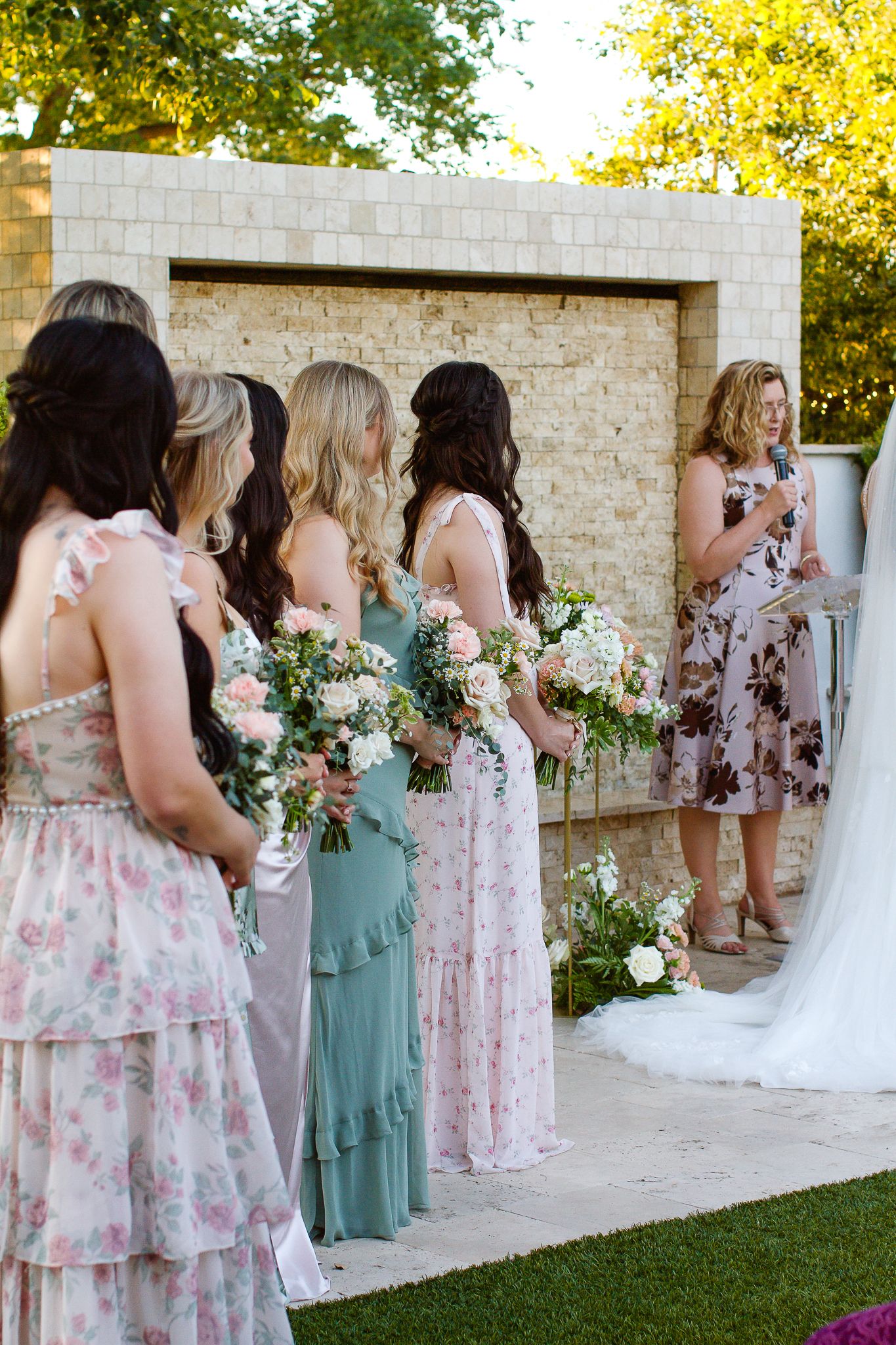 Wedding party members in floral and sage green dresses stand in a row along a stone colonnade during an outdoor ceremony.