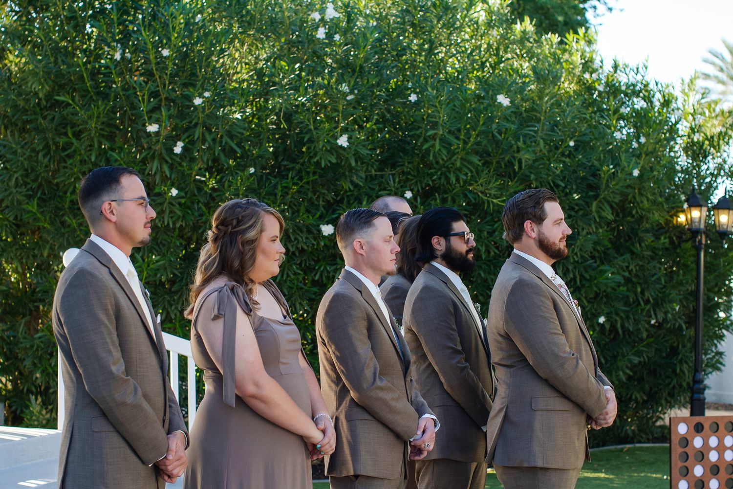 Wedding party poses outdoors in brown and gray formal attire during sunset at a garden venue.