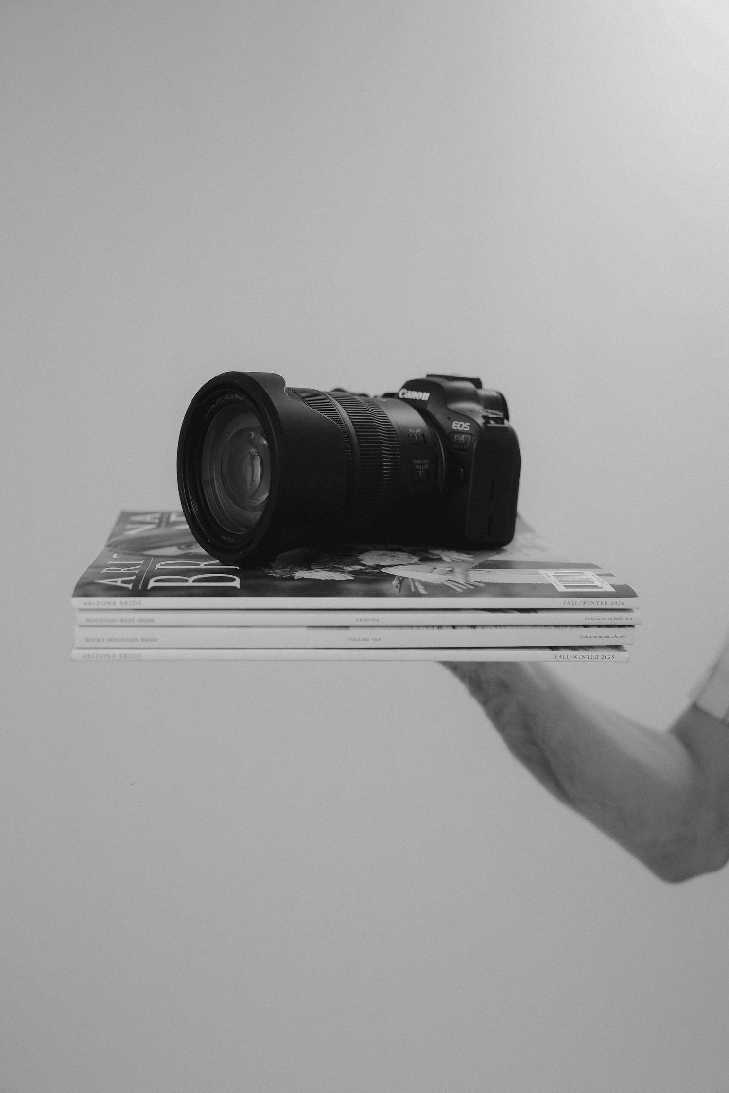 Black and white photo of a DSLR camera with wide-angle lens resting on stacked magazines held by a hand.