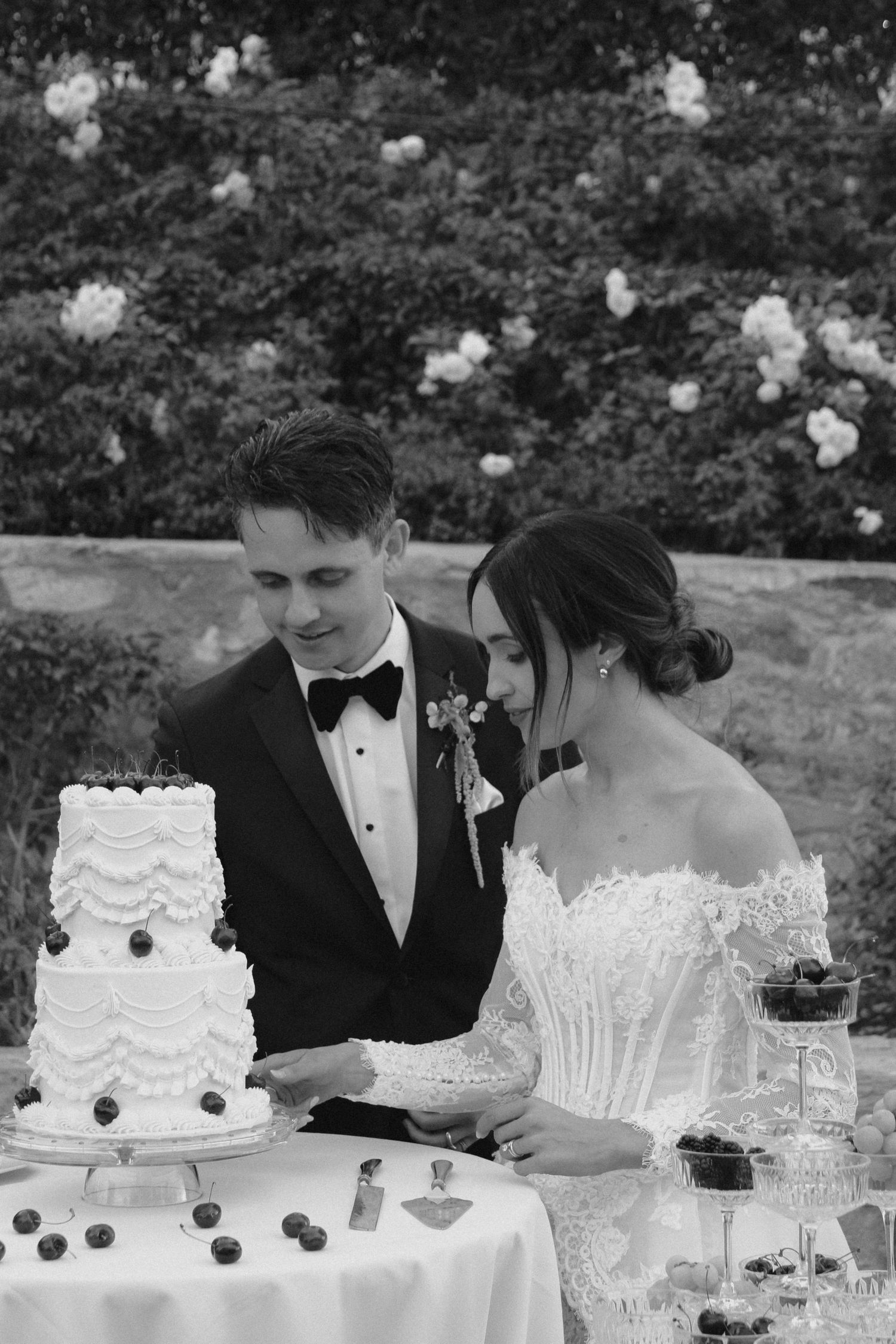 Black and white photo of bride and groom cutting their tiered wedding cake outdoors surrounded by blooming flowers.