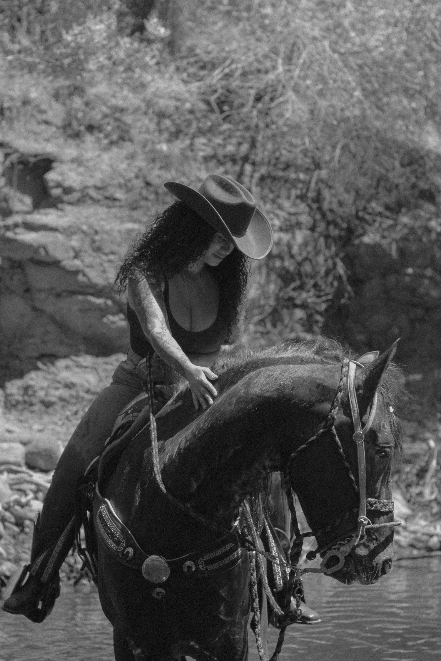 A rider in western attire sits atop a dark horse near a water's edge in a black and white photograph.
