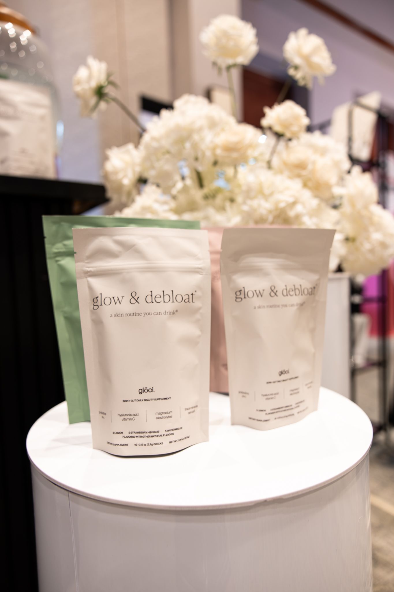 Glow & Debloat supplement pouches in green and pink displayed on white pedestal with white flowers.