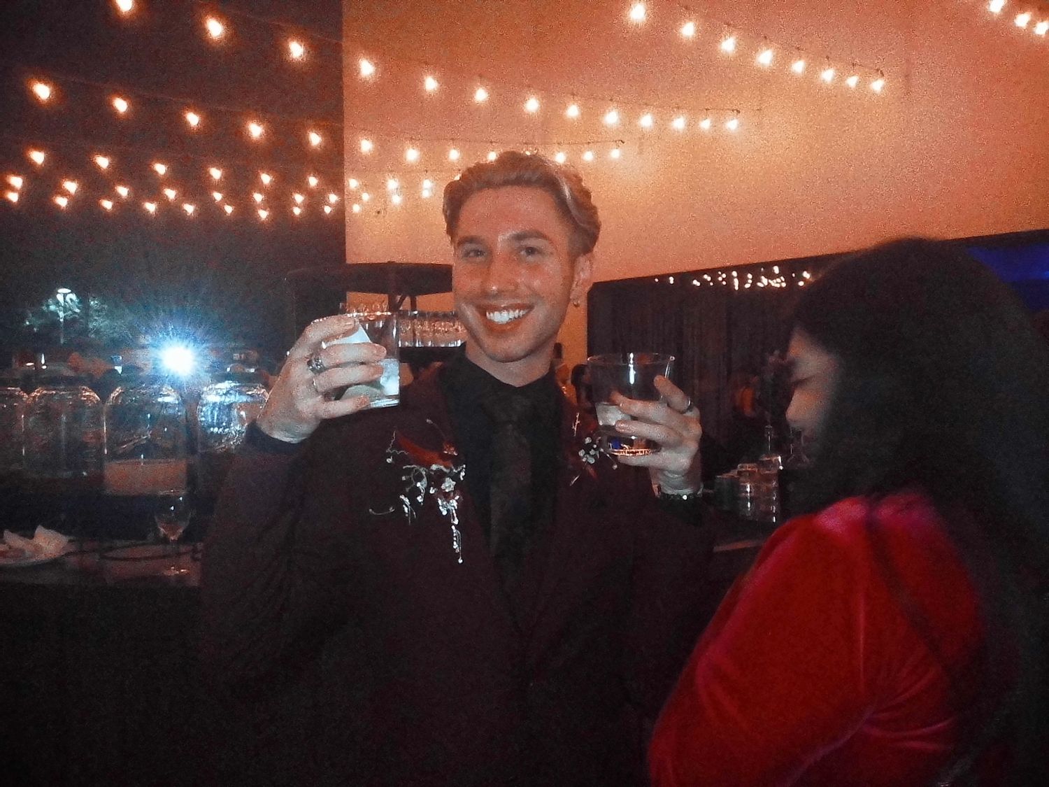 Smiling young man in a black jacket raising a glass at an outdoor evening event with string lights.
