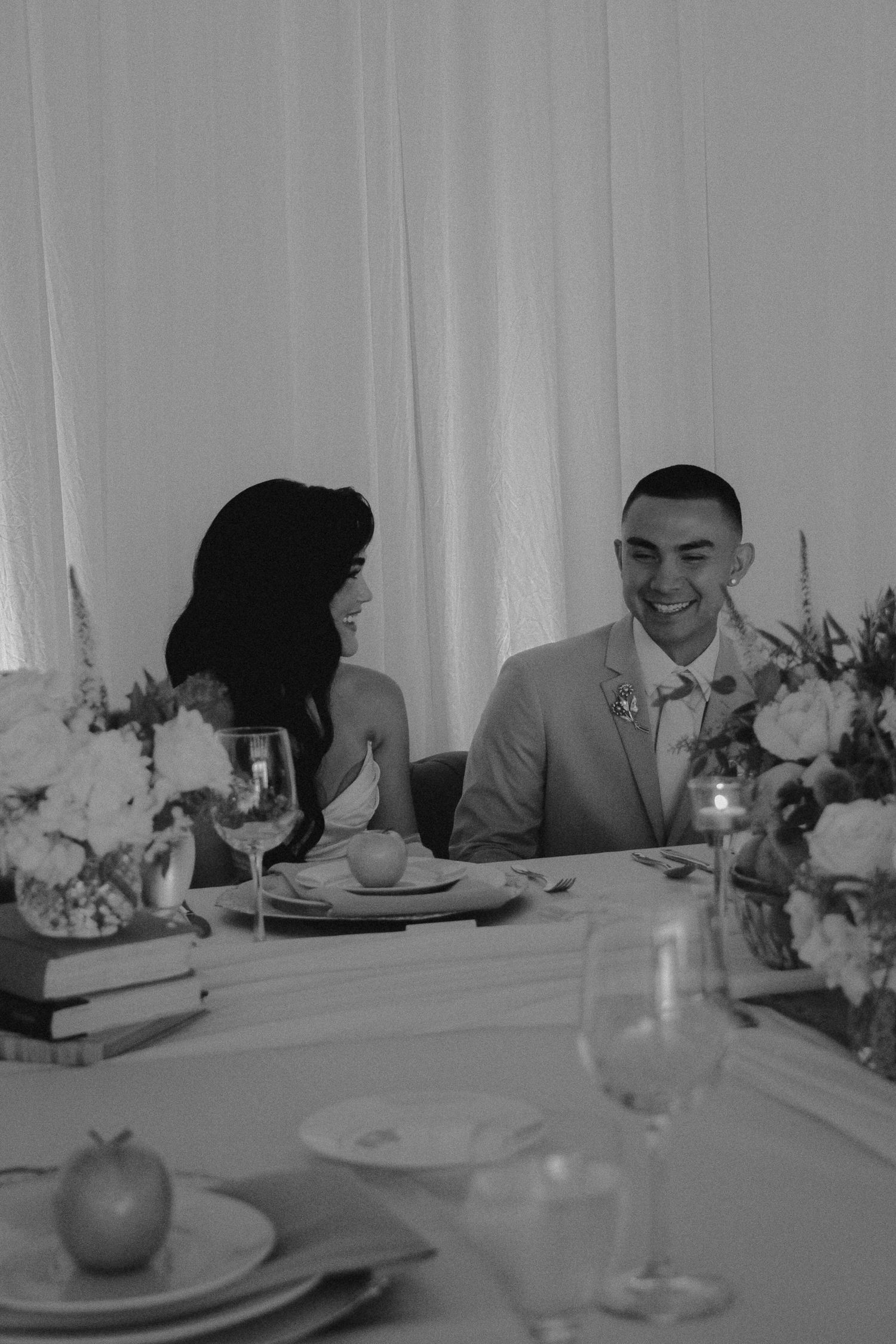 Bride and groom laughing together at sweetheart table with floral centerpieces at wedding reception.