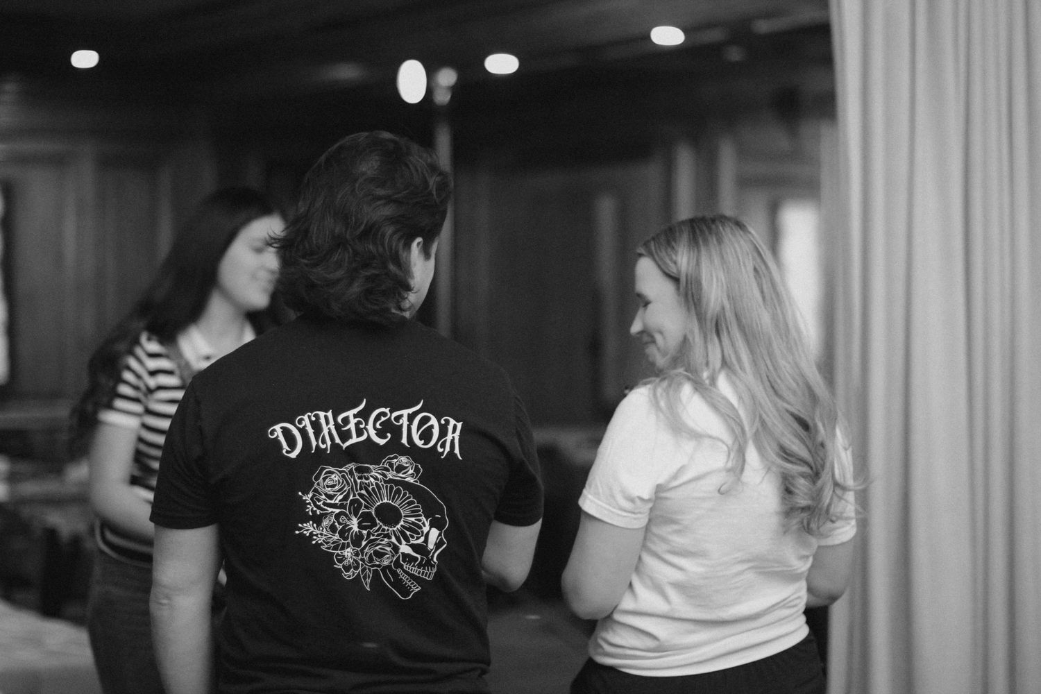 Black and white photo of three people talking, one wearing a Director t-shirt with skull graphic design.