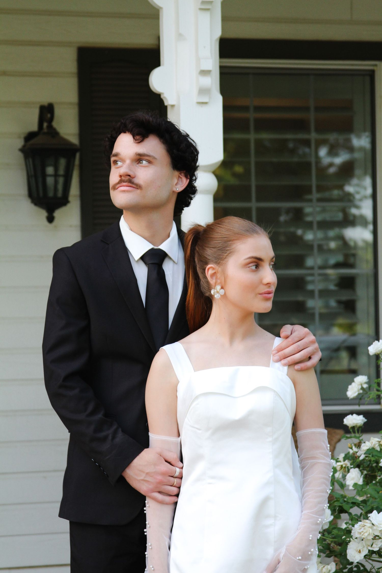 A couple in wedding attire poses outside a house, groom in black suit and bride in white dress with gloves.
