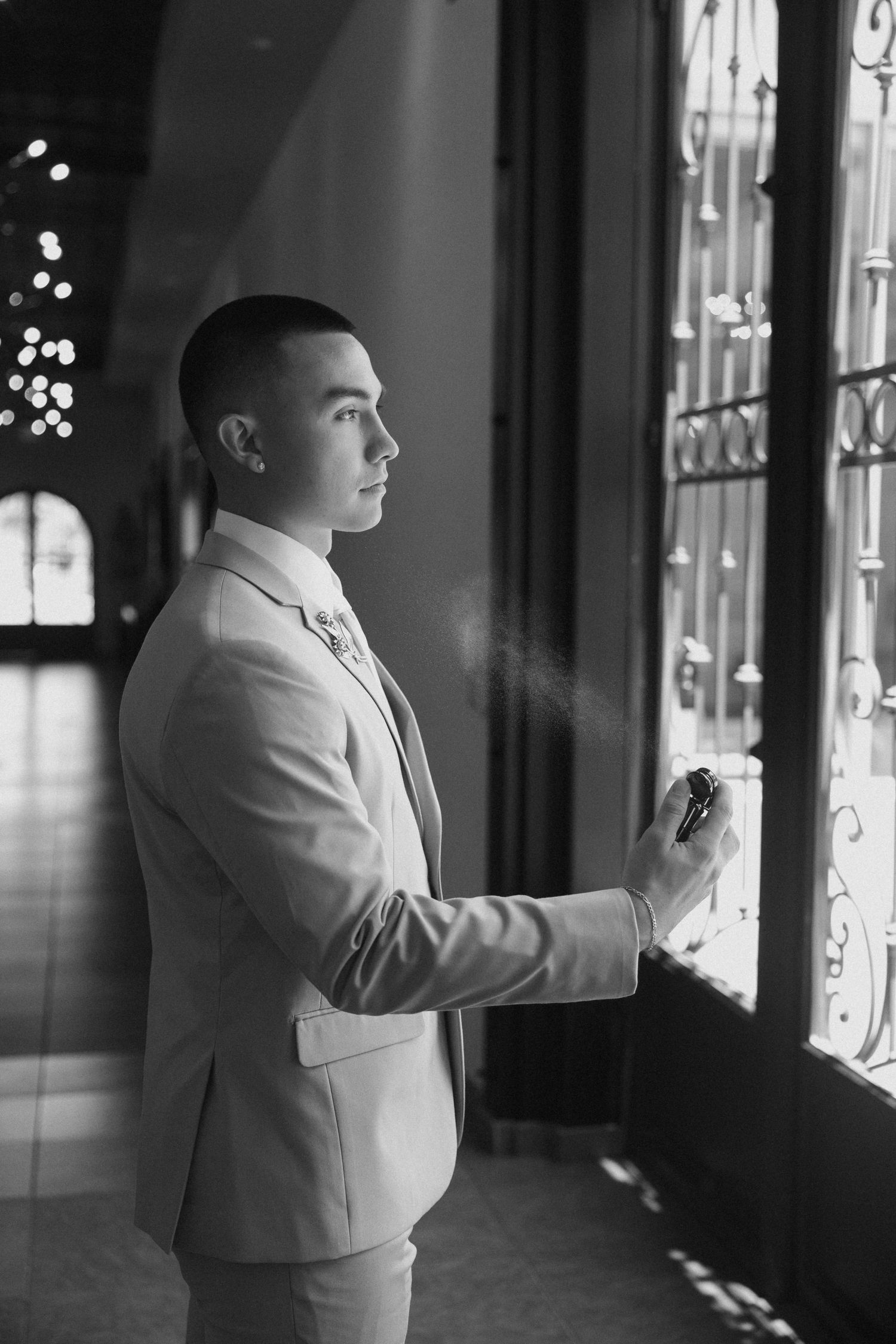 Black and white portrait of groom in tan suit spraying cologne near ornate window before wedding.