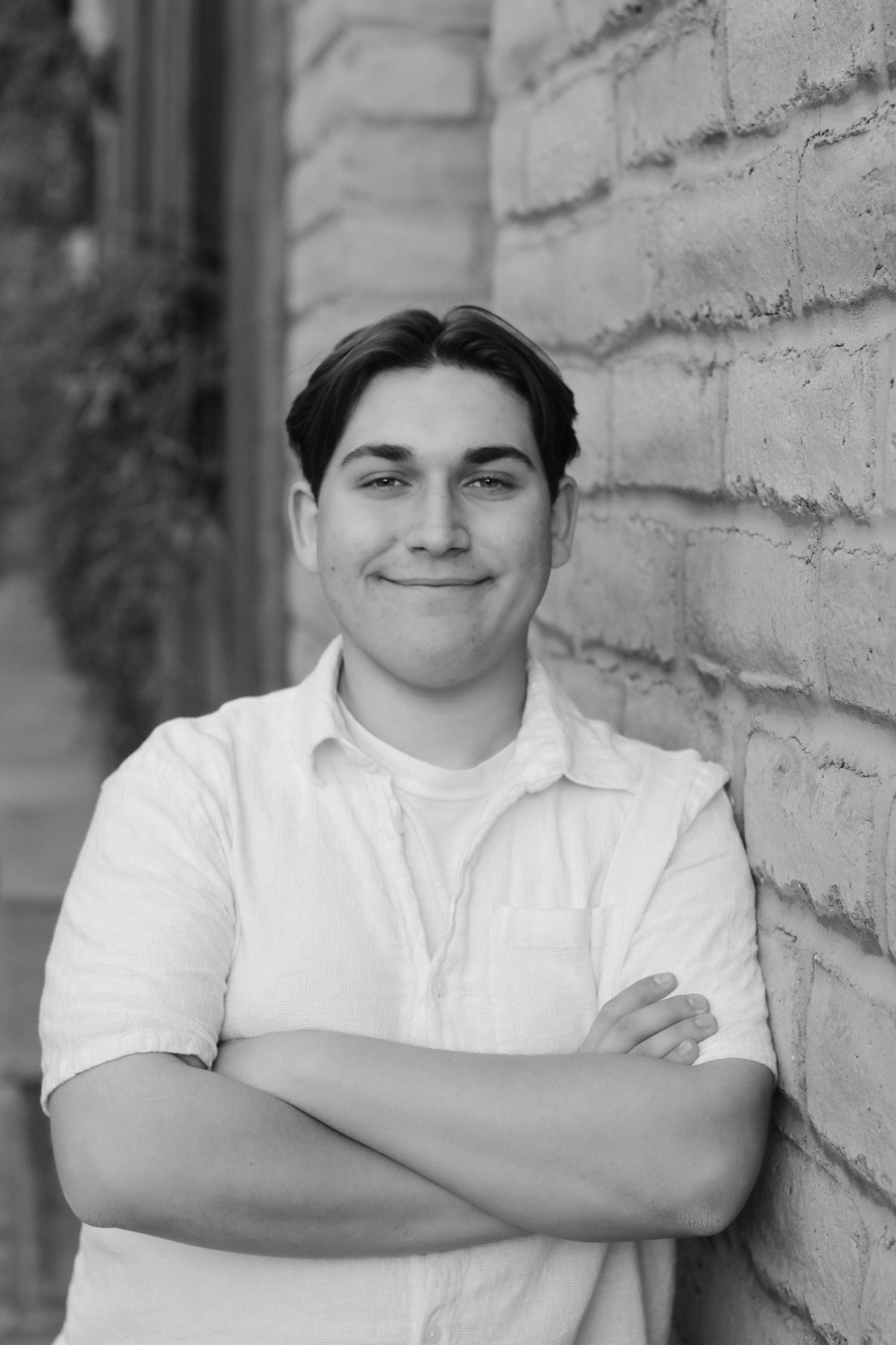 Graduate in white shirt standing with arms crossed against textured brick wall in black and white portrait.