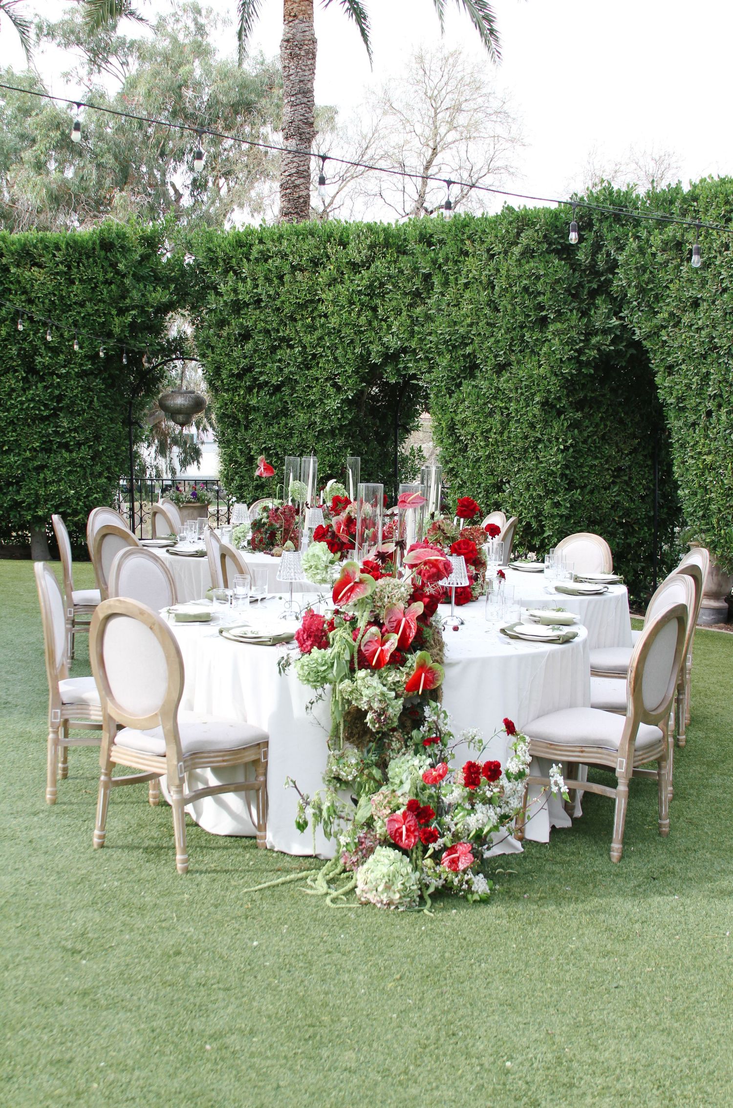 Elegant outdoor wedding reception table with red floral centerpiece, white linens, and gold Louis chairs.